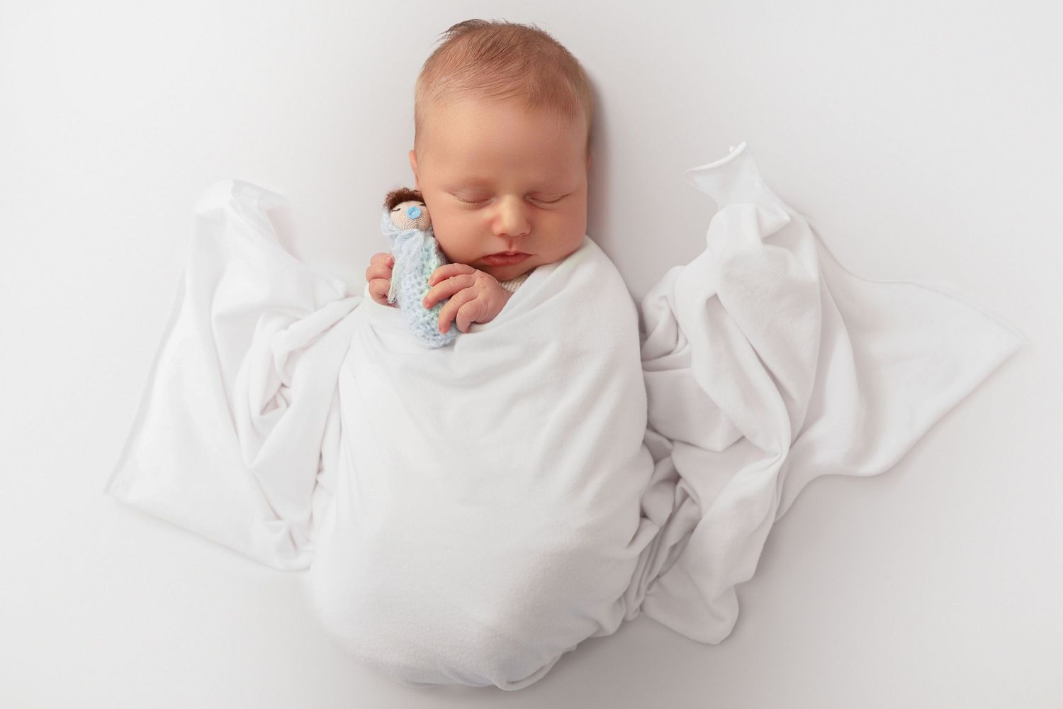 Peaceful newborn baby swaddled in white blanket sleeping with a small blue toy on a white background.
