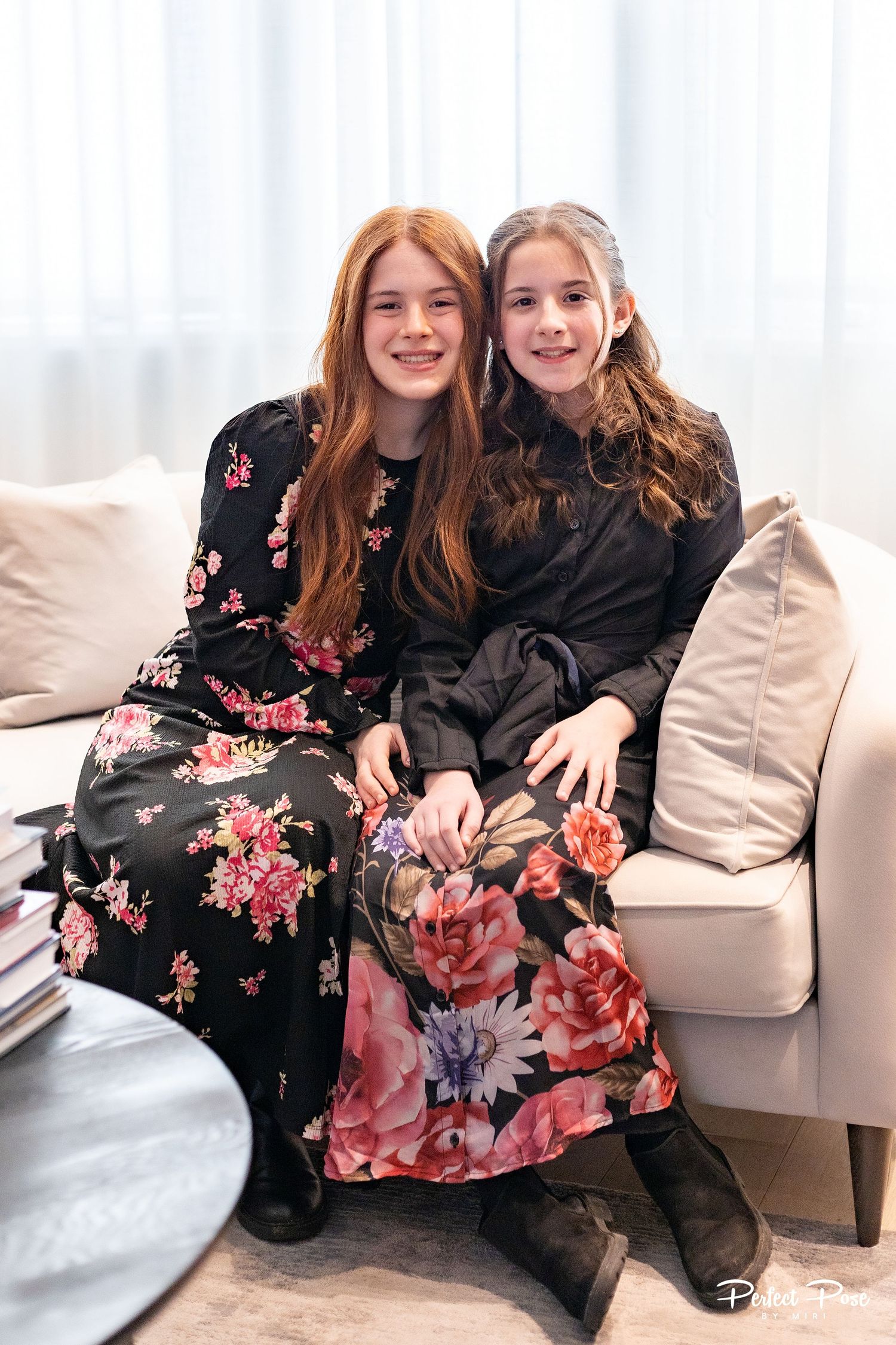 Two young girls in floral dresses sitting together on a white sofa, smiling warmly in a bright modern room.