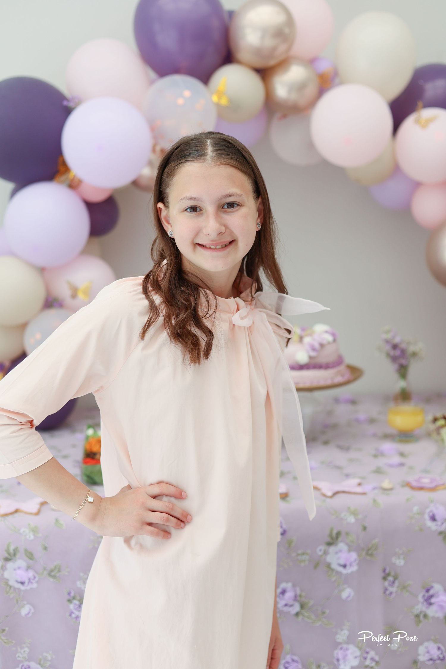 A smiling girl in a pink dress poses at a purple-themed birthday party with balloon arch and decorated cake table.