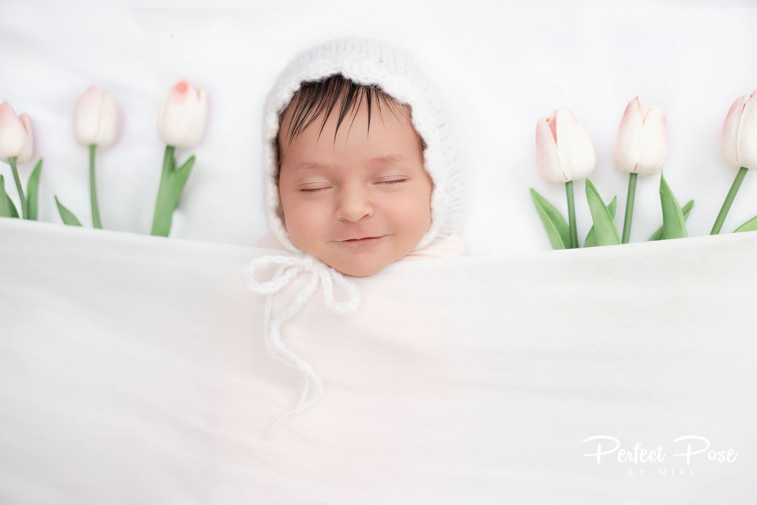 Newborn baby sleeping peacefully wrapped in white, wearing a knit bonnet, surrounded by pink tulips.