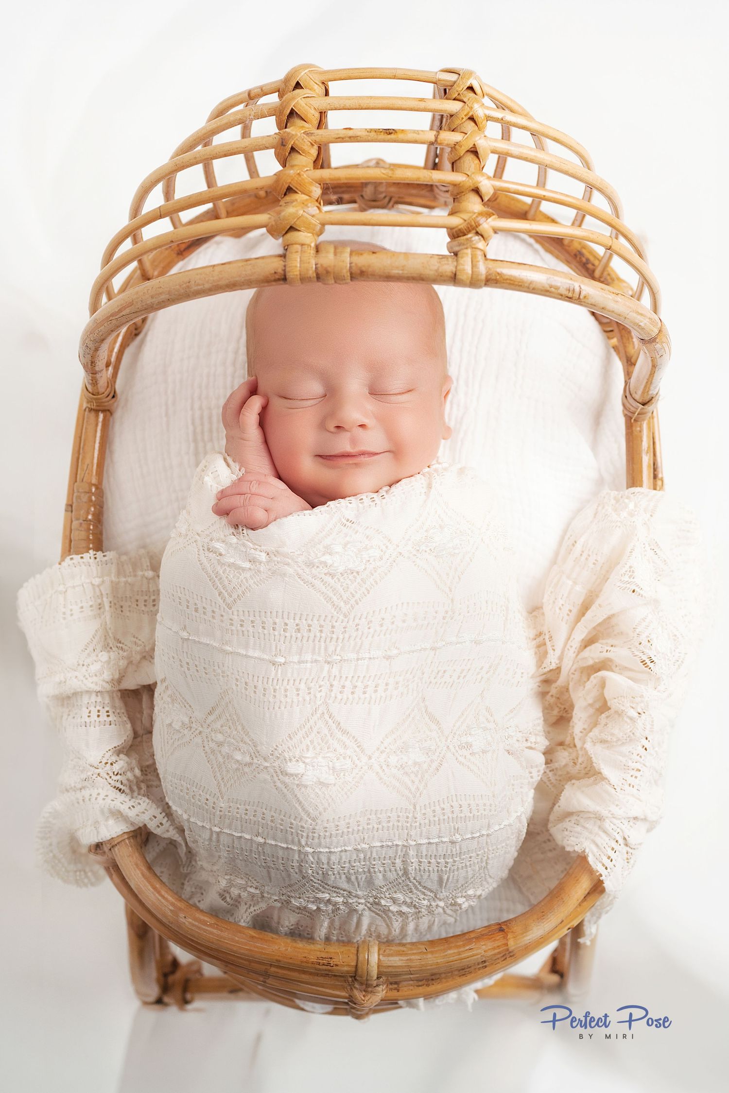 Smiling newborn baby swaddled in cream knit wrap, sleeping peacefully in a small rattan bassinet on white background.