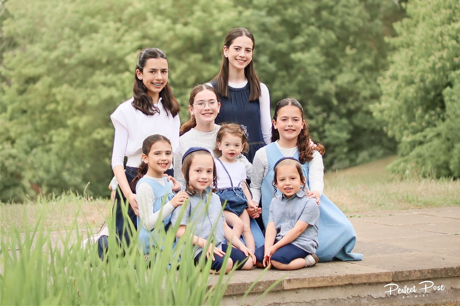 Family portrait of two women and six young girls in matching blue dresses posing outdoors in a lush green park.