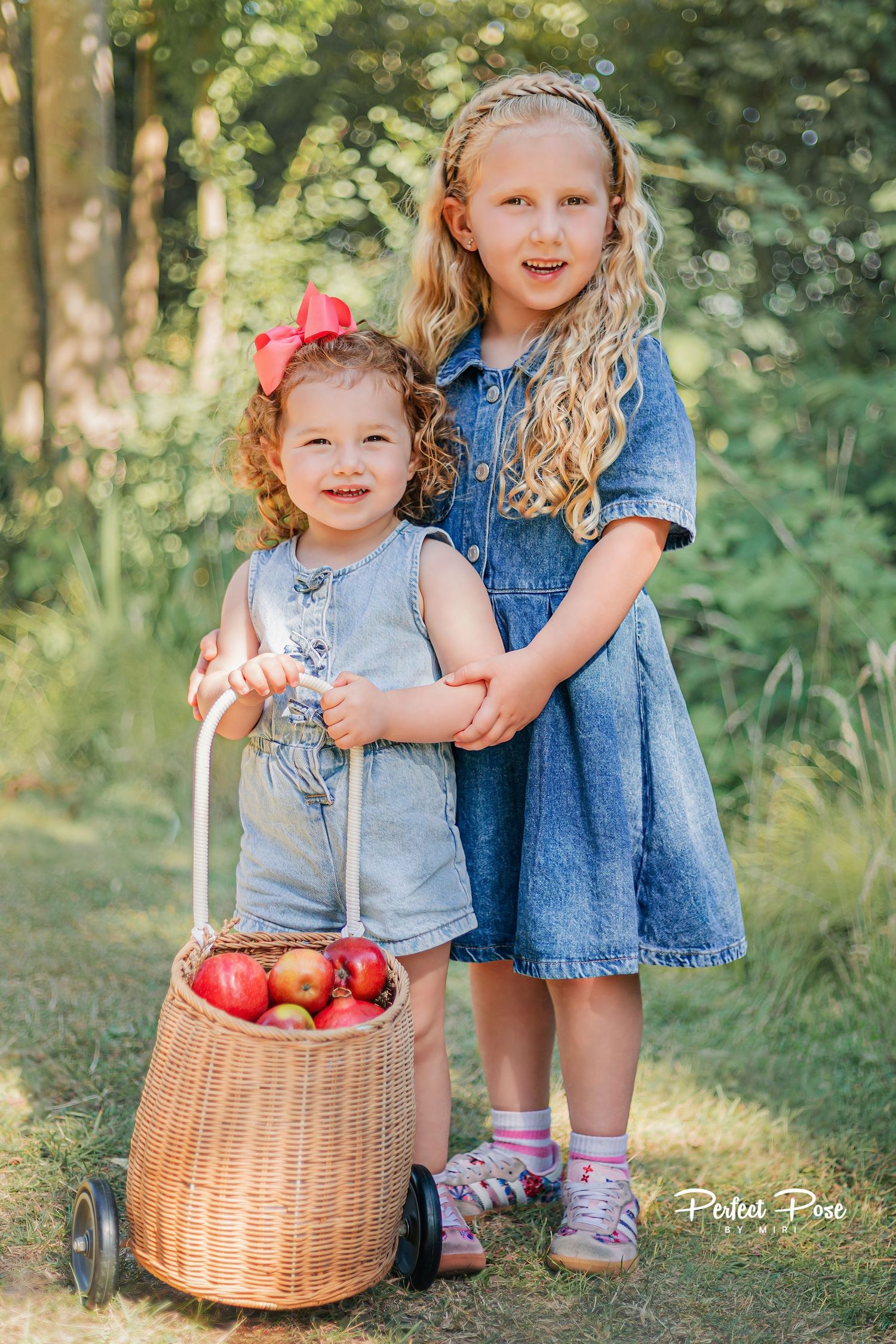 Two young sisters in denim outfits pose outdoors with a wicker basket cart full of red apples.