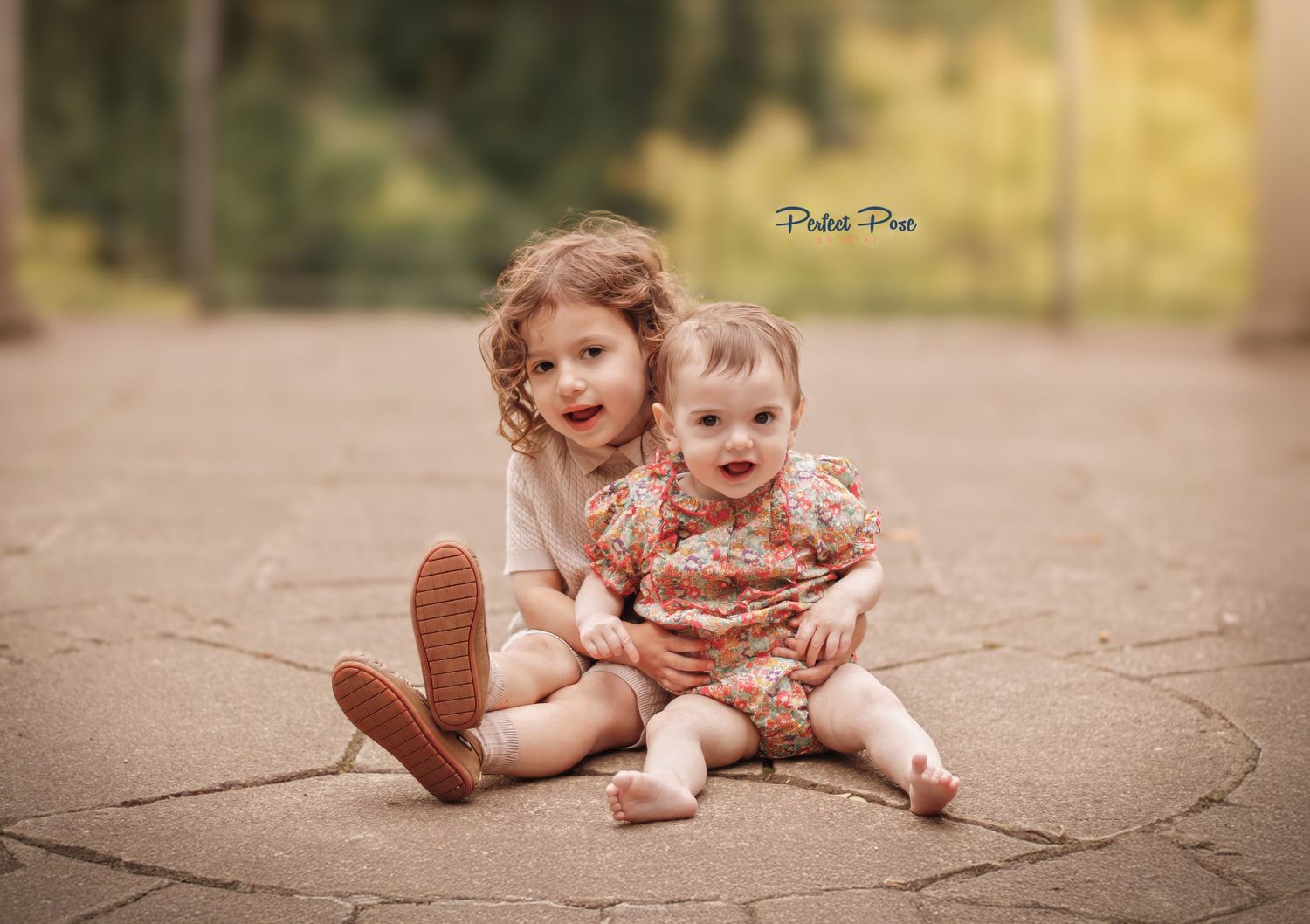 Two young siblings sit together on a path, older toddler hugging baby in floral outfit, outdoor portrait photography.