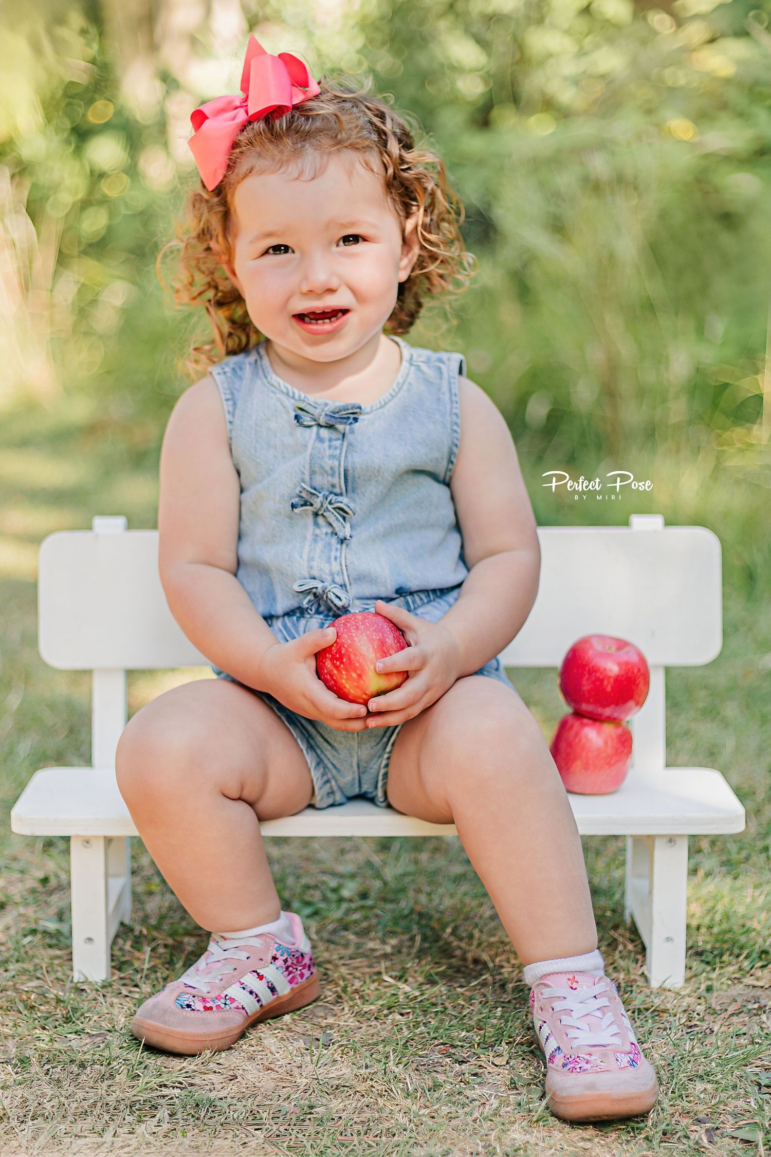 Toddler girl with curly hair and red bow sits on white bench holding red apples outdoors.