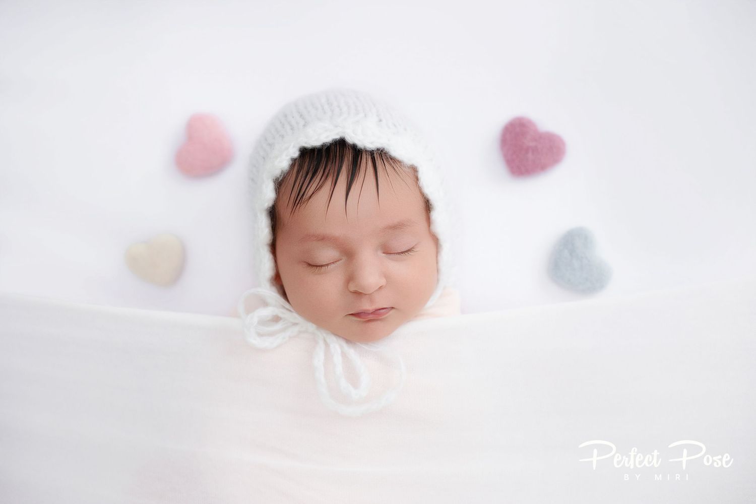 Sleeping newborn baby wearing a white knit bonnet surrounded by colorful heart props on a white background.