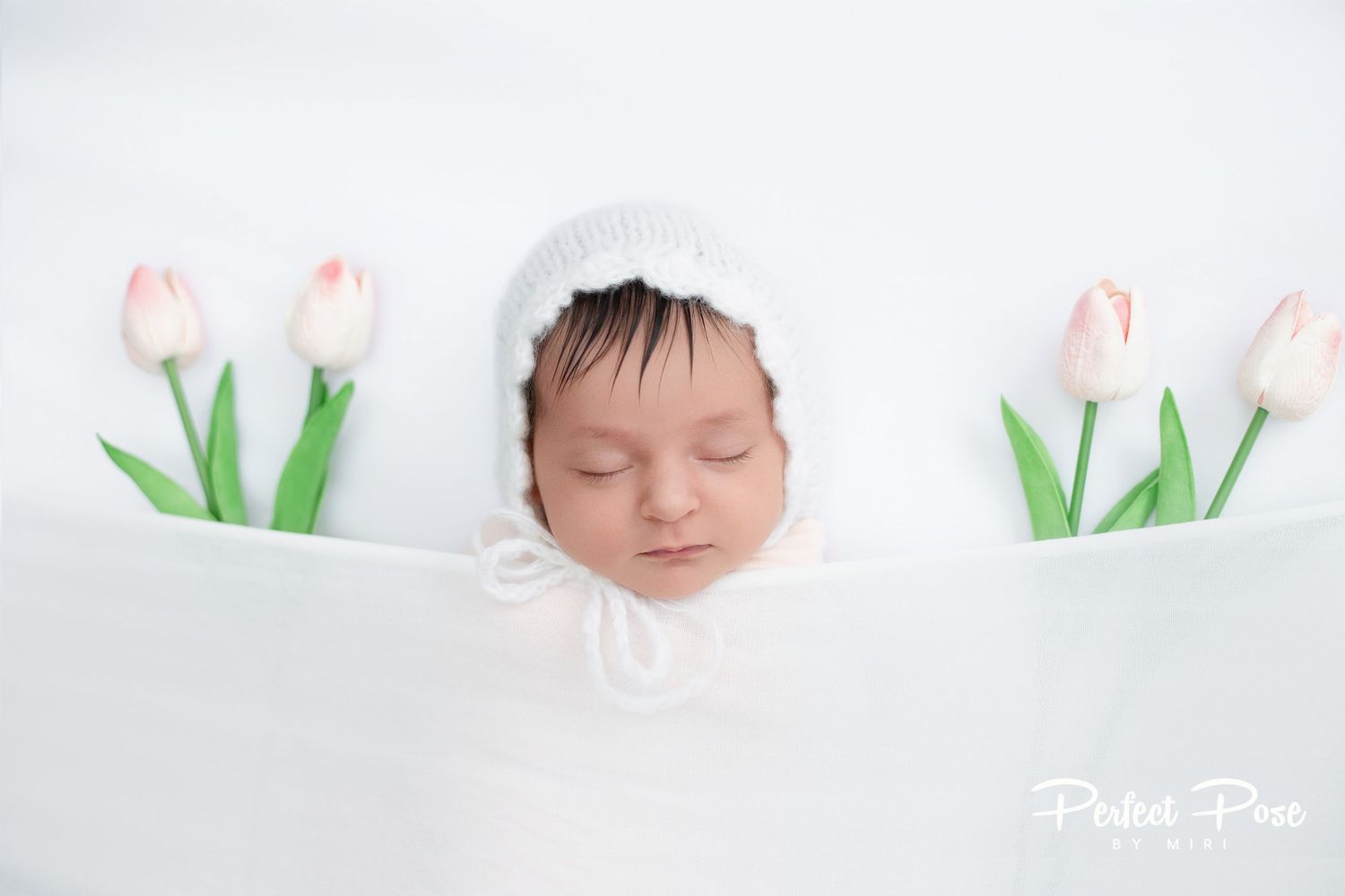 Sleeping newborn baby in white bonnet surrounded by pink tulips in a soft white wrap, professional newborn photography.
