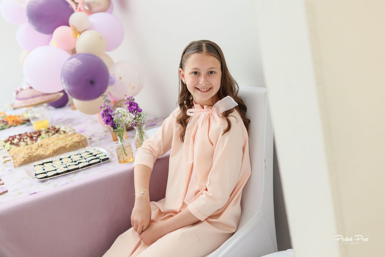A smiling girl in a pink outfit sits at a decorated birthday party table with balloons and treats.