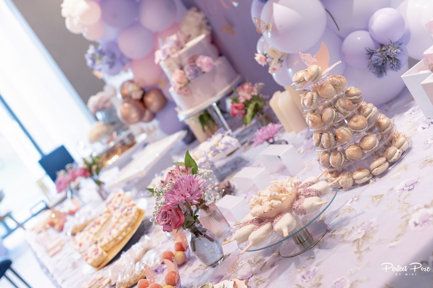 Elegant dessert table with macarons, cakes, and floral decor under a lavender balloon arch at a party.