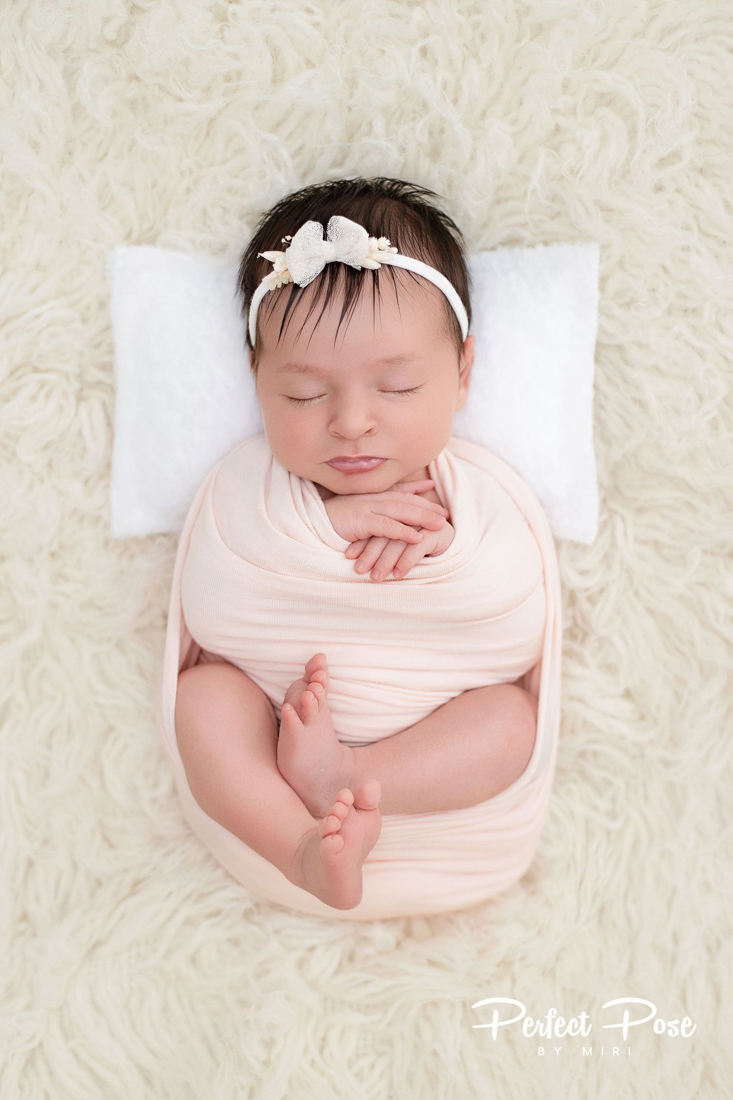 Sleeping newborn baby girl swaddled in pink wrap with floral headband on cream fur backdrop.