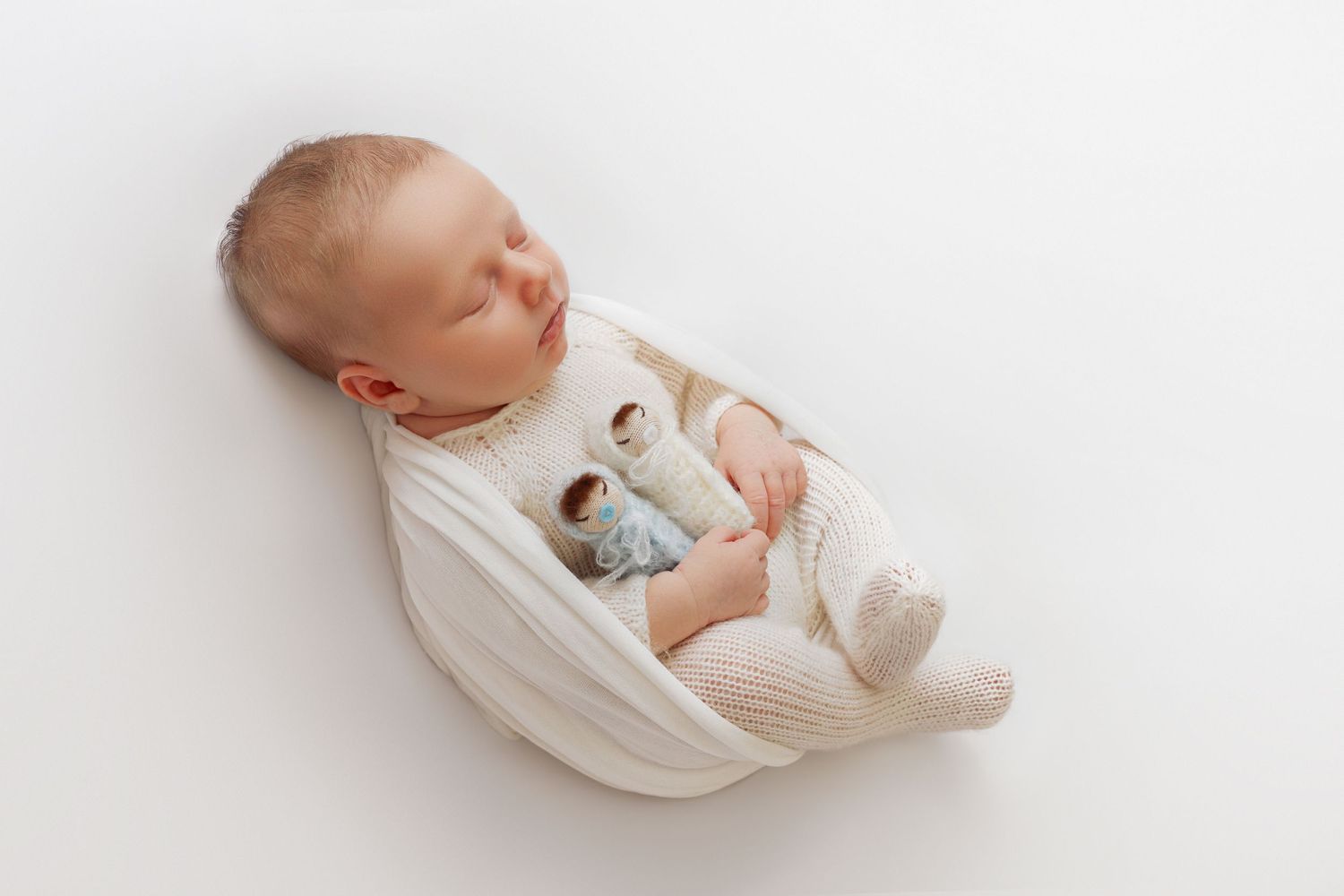 Sleeping newborn baby wrapped in cream knit swaddle holding a tiny doll toy on white background.