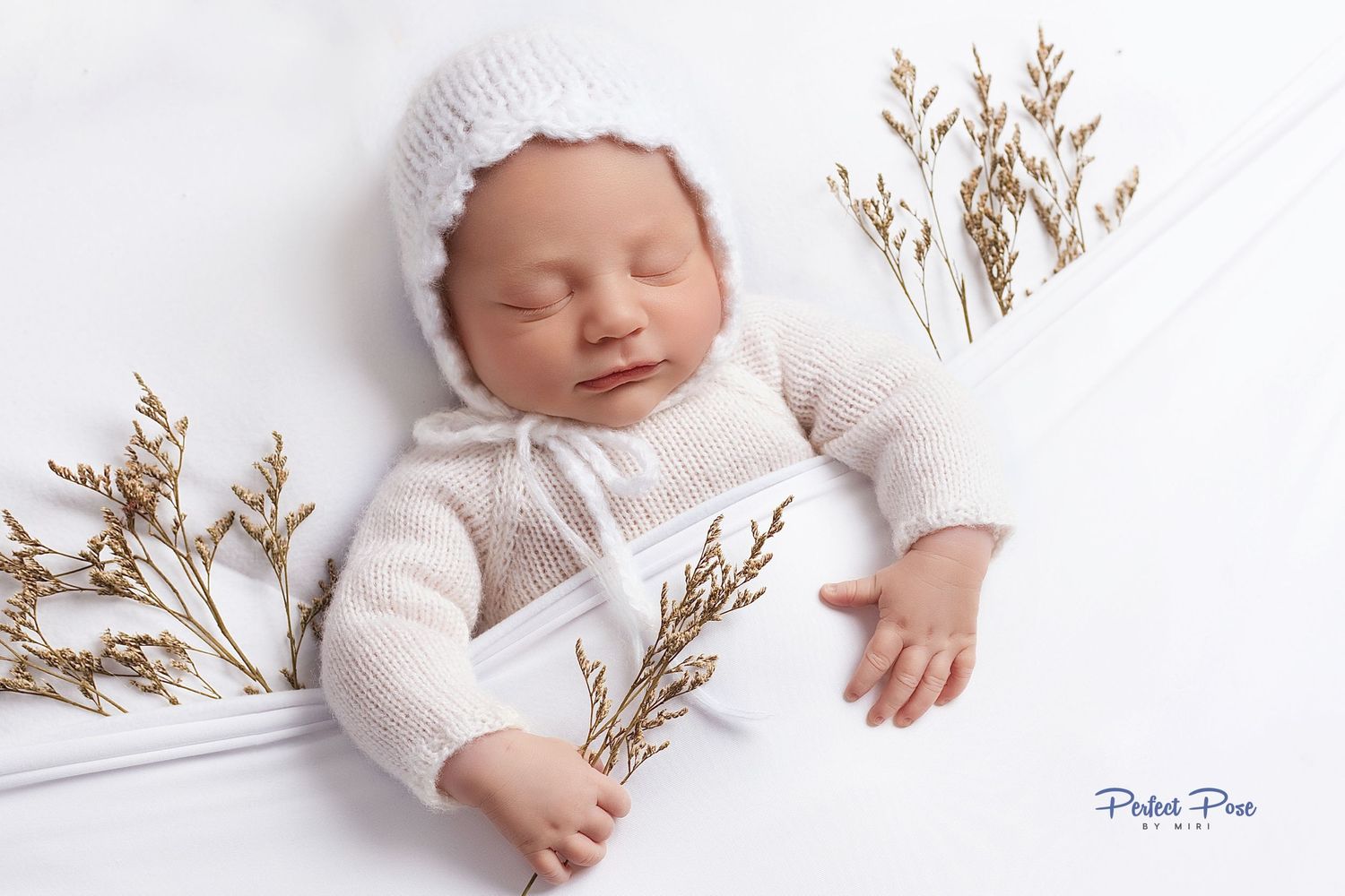 Sleeping newborn baby in white knit romper and bonnet surrounded by golden dried flowers on white background.