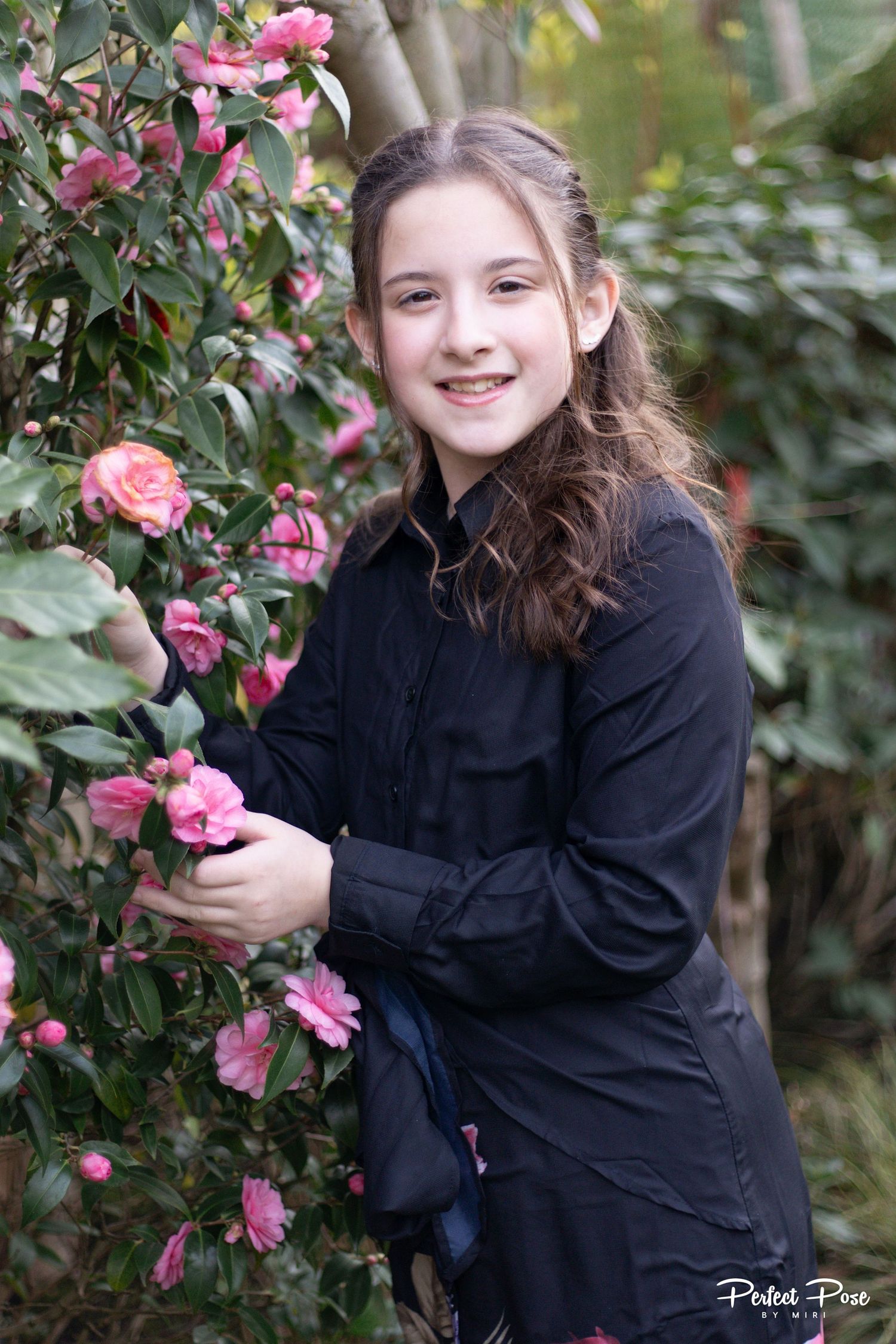Young girl in black dress smiling while holding pink camellia flowers in a garden setting.