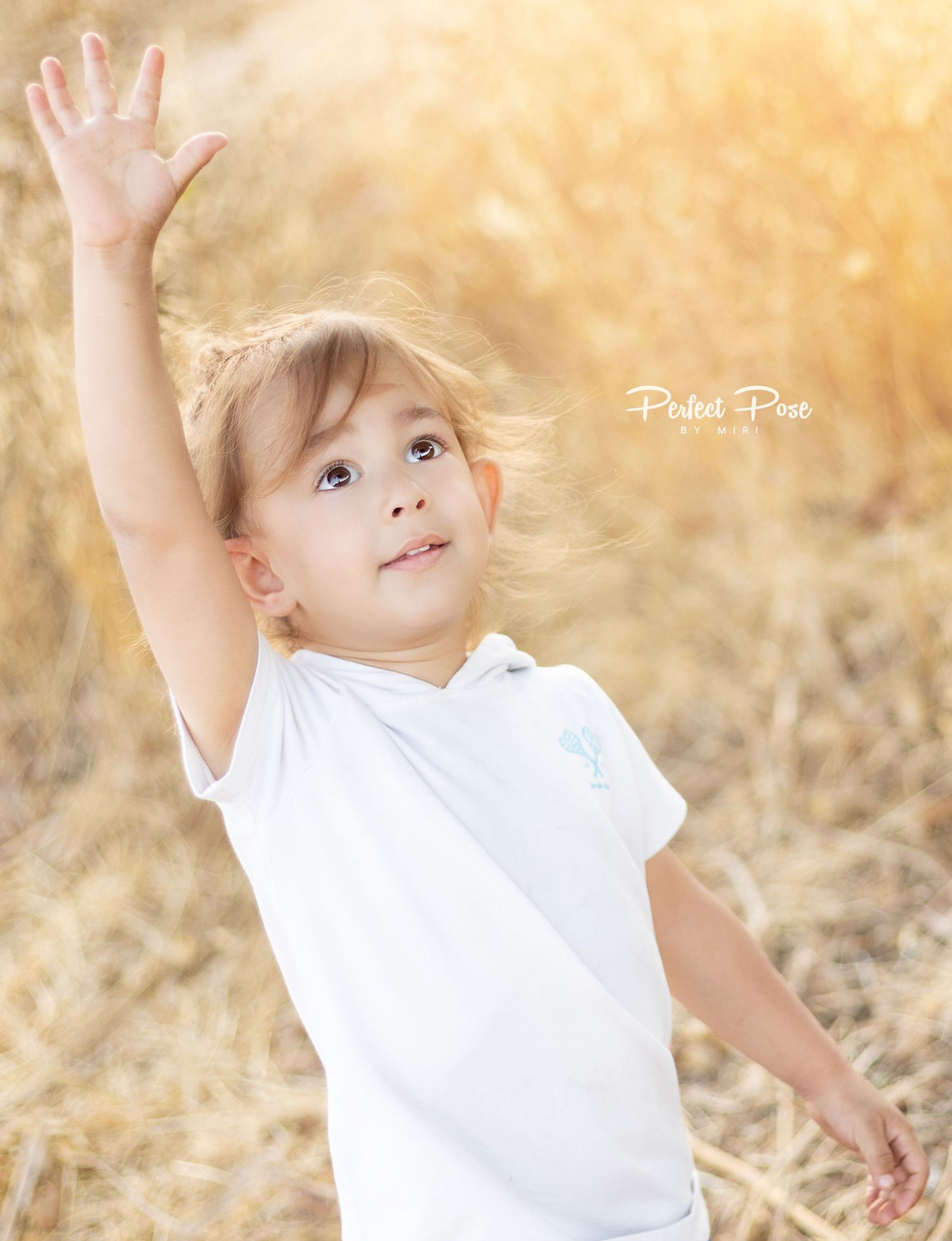 A joyful toddler in a white shirt raises her hand up outdoors in a golden sunlit field.
