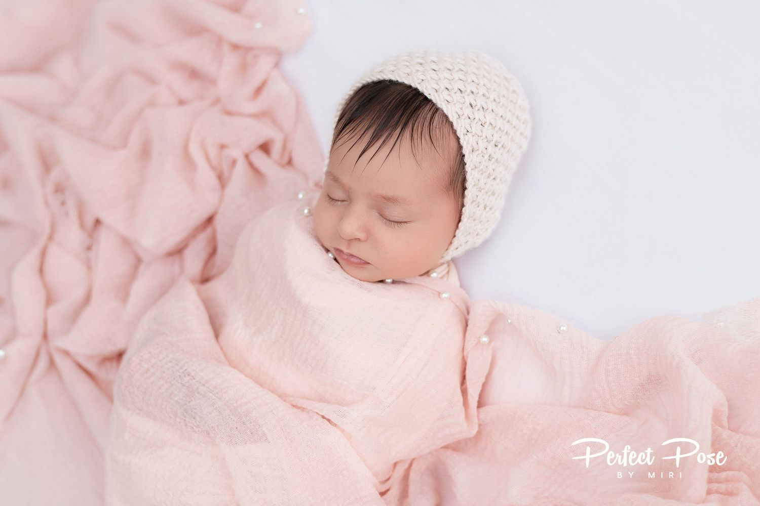 Newborn baby sleeping peacefully wrapped in pink muslin with a cream knit bonnet on white backdrop.