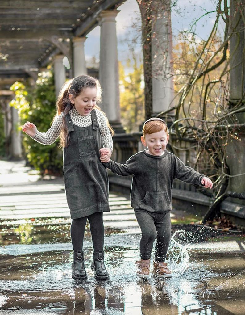 Two happy children splashing in puddles outdoors, wearing coordinating dark outfits and boots on a sunny autumn day.