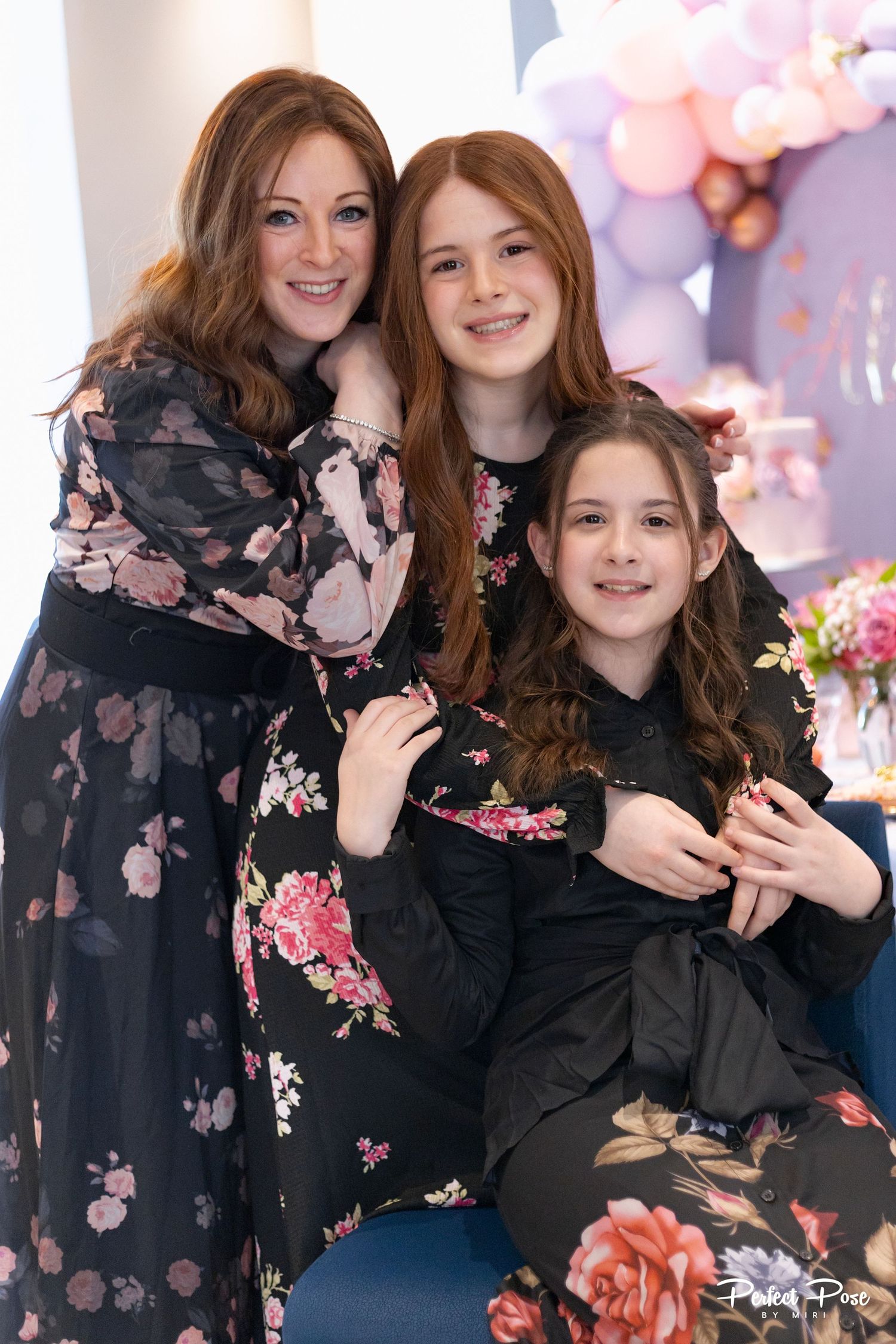 A woman and two red-haired girls in floral dresses smile together at a party with pink balloon decor.
