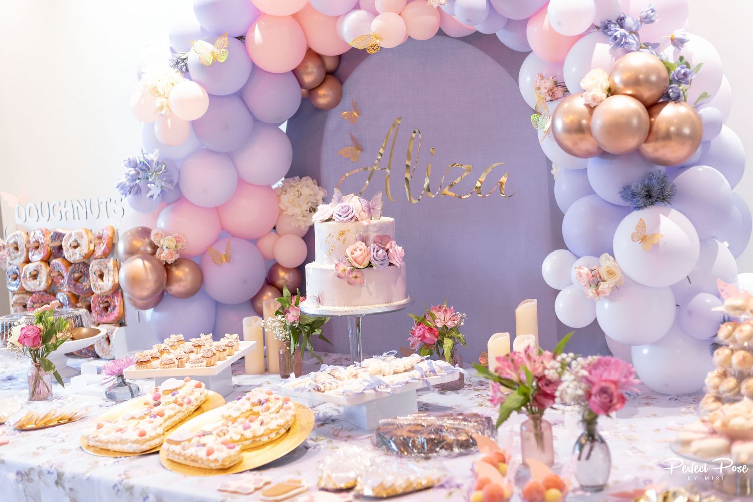 Elegant birthday dessert table with pastel balloon arch, floral cake, and assorted sweets in pink, lavender, and gold tones.