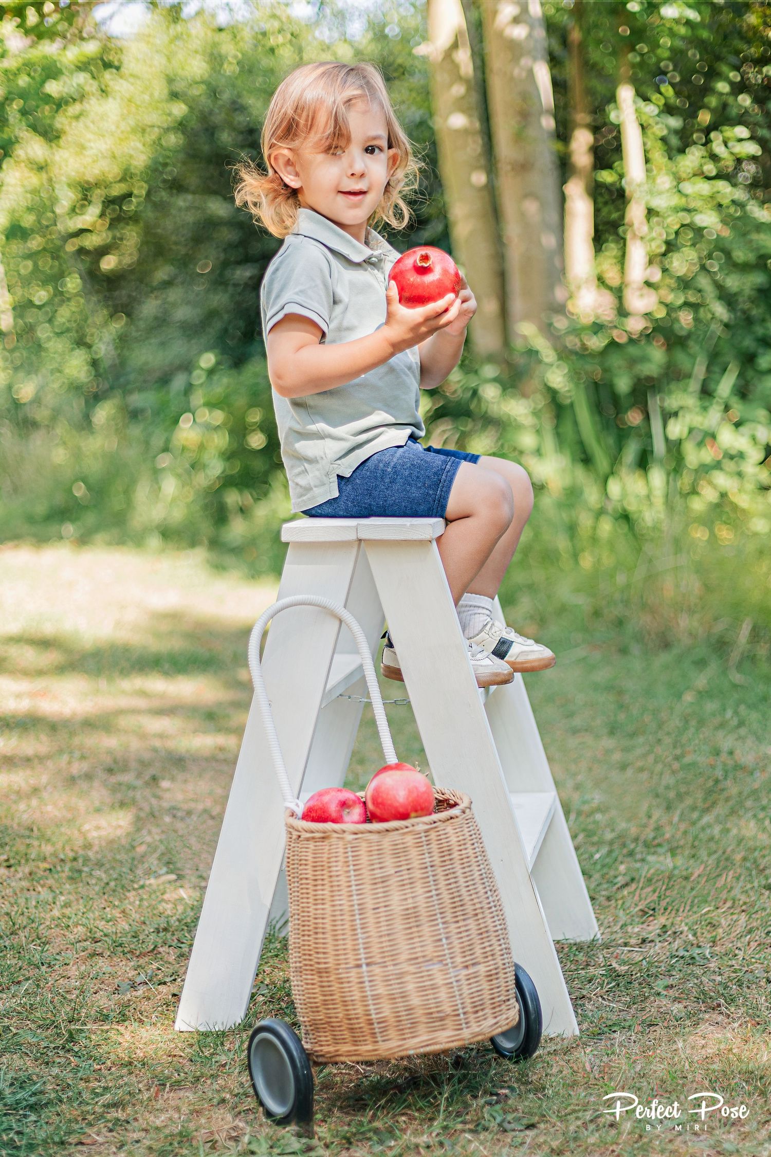 A young child sits on a white step stool holding a red apple, with a wicker basket of apples below, outdoors.