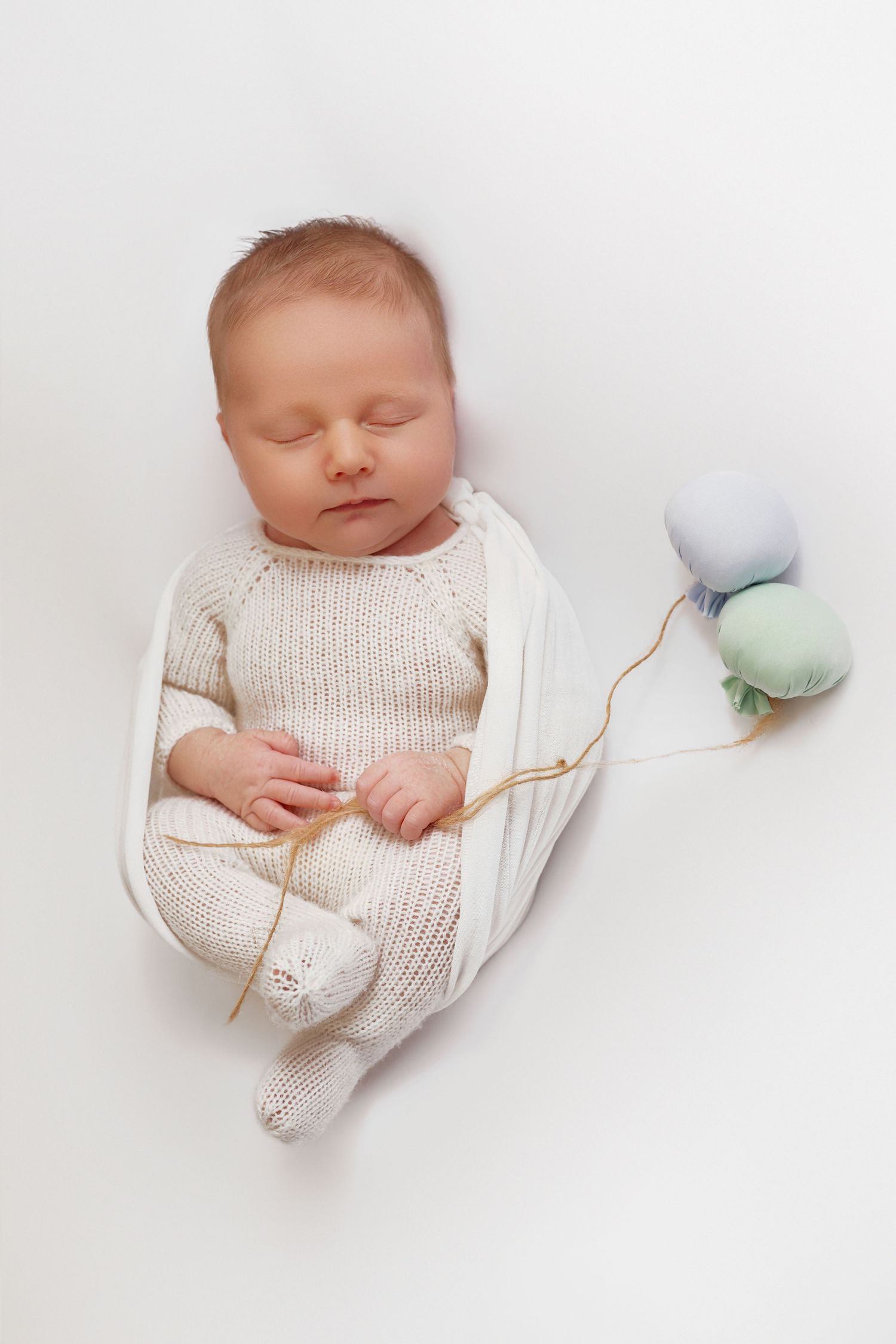 Sleeping newborn baby in cream knit outfit wrapped in white blanket holding pastel yarn balls on white background.