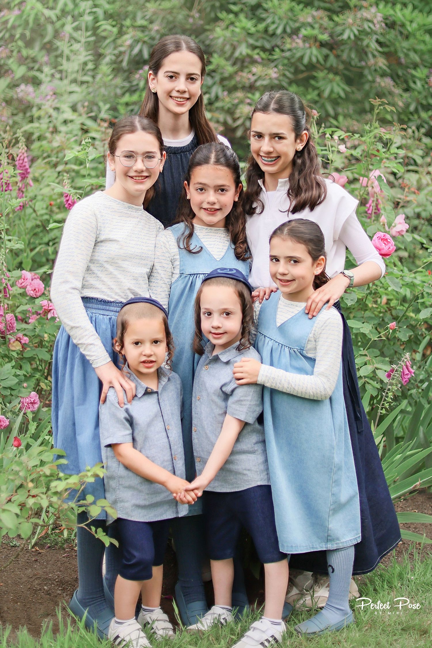 Group of seven girls posing together in a garden with pink flowers, wearing coordinated blue and gray outfits.