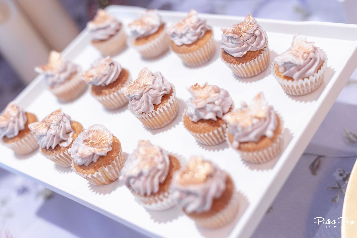 Elegant cupcakes with lavender frosting and gold leaf decorations arranged on a white tray at a wedding reception.