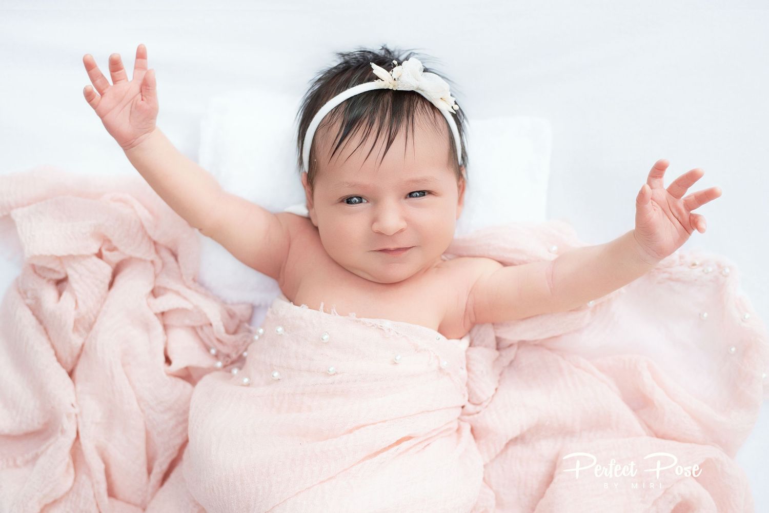 Adorable baby girl in pink wrap with white bow headband, arms raised joyfully during newborn photo session.