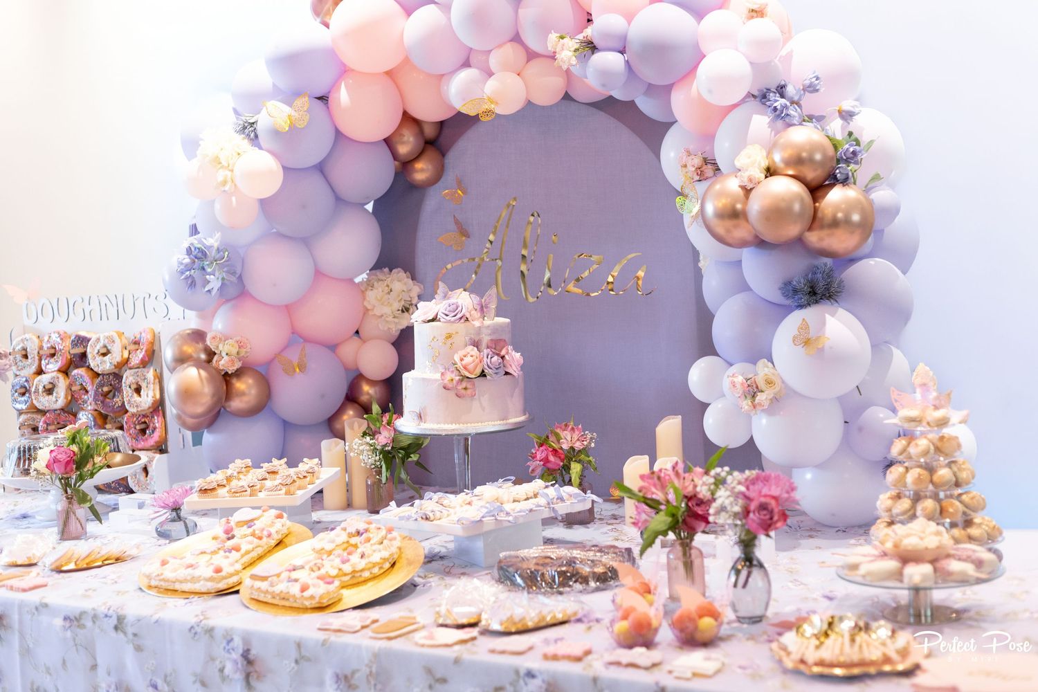 Elegant dessert table with pastel balloon arch, floral cake, donuts, and sweets in pink, lavender, and gold tones.