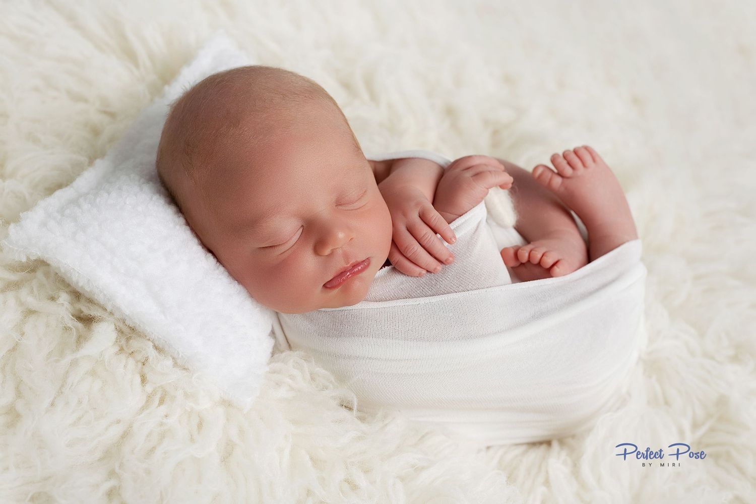 Peaceful newborn baby swaddled in white, sleeping on a fluffy cream blanket with a tiny white pillow.