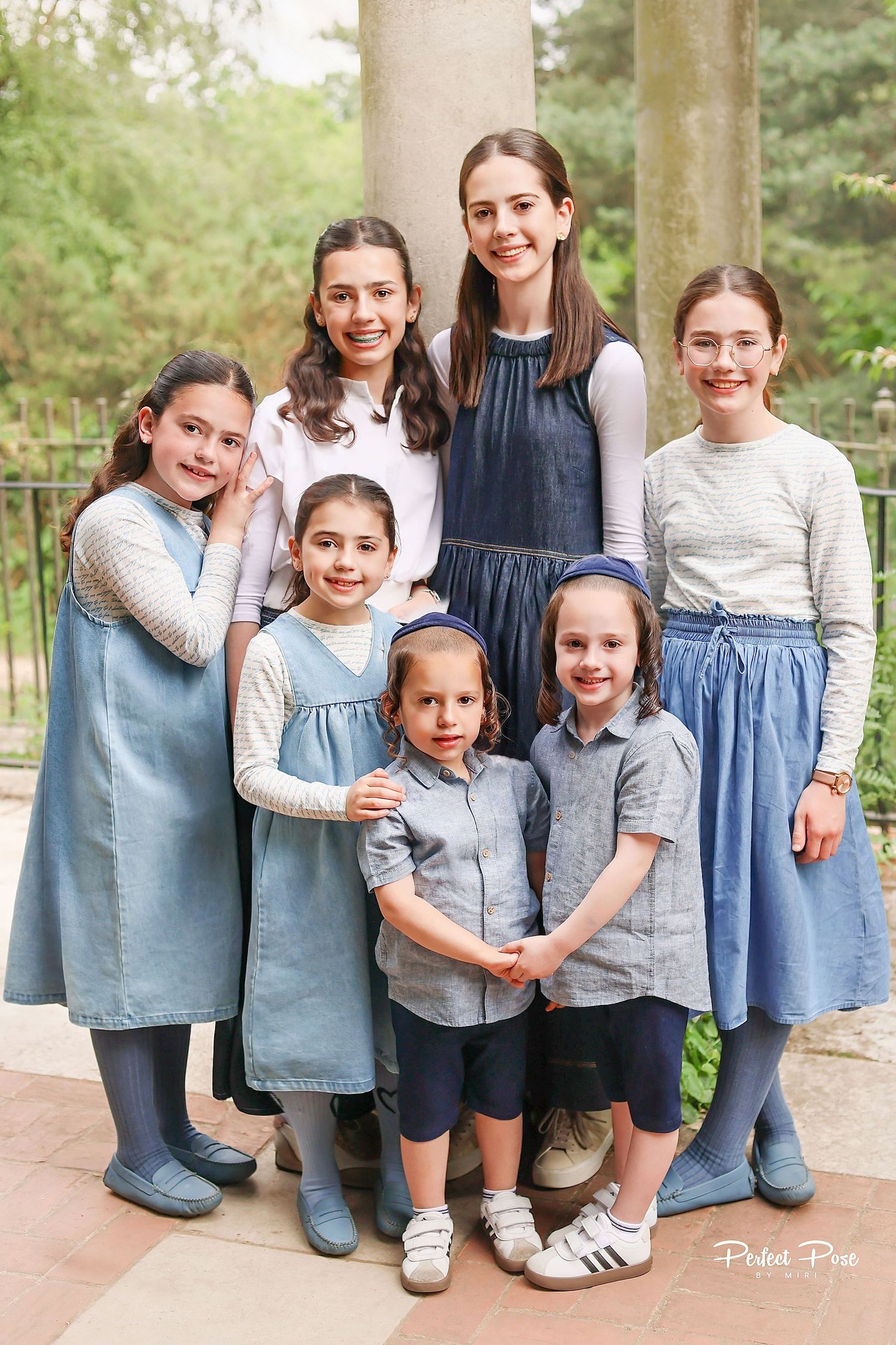 Group of seven girls in modest blue dresses and skirts posing together outdoors near stone columns.