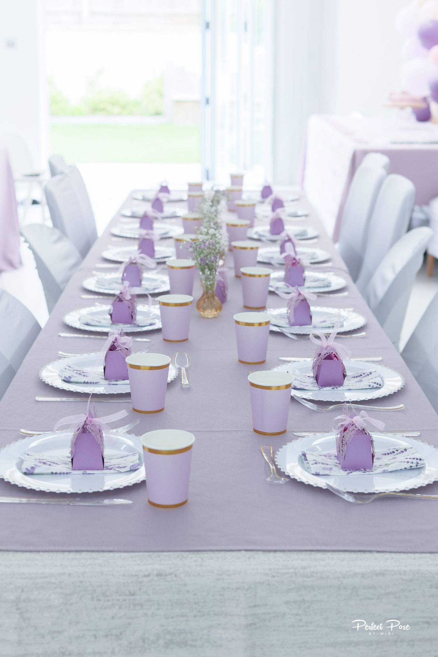 Elegant lavender party table setting with purple cups, decorative favor boxes, and silver charger plates for a celebration.
