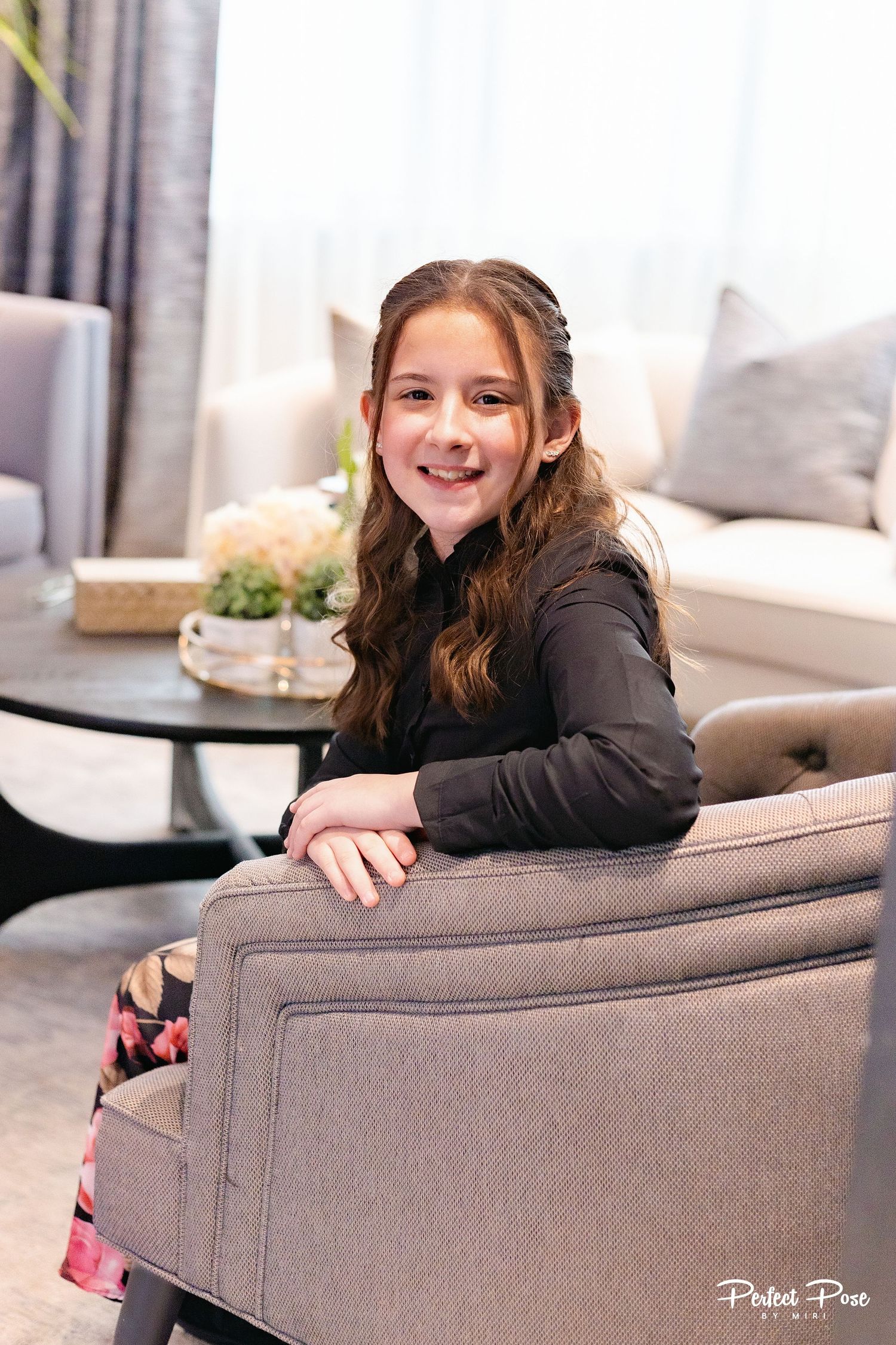 A smiling young girl with long wavy hair sits in a gray armchair in an elegant living room.