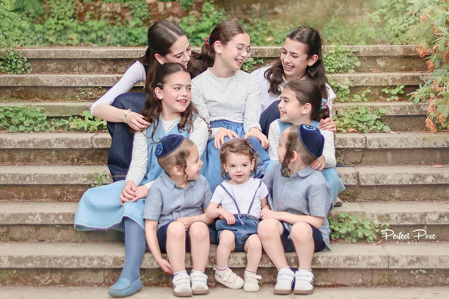 Group of children in matching blue outfits sitting together on stone steps, smiling and hugging outdoors.