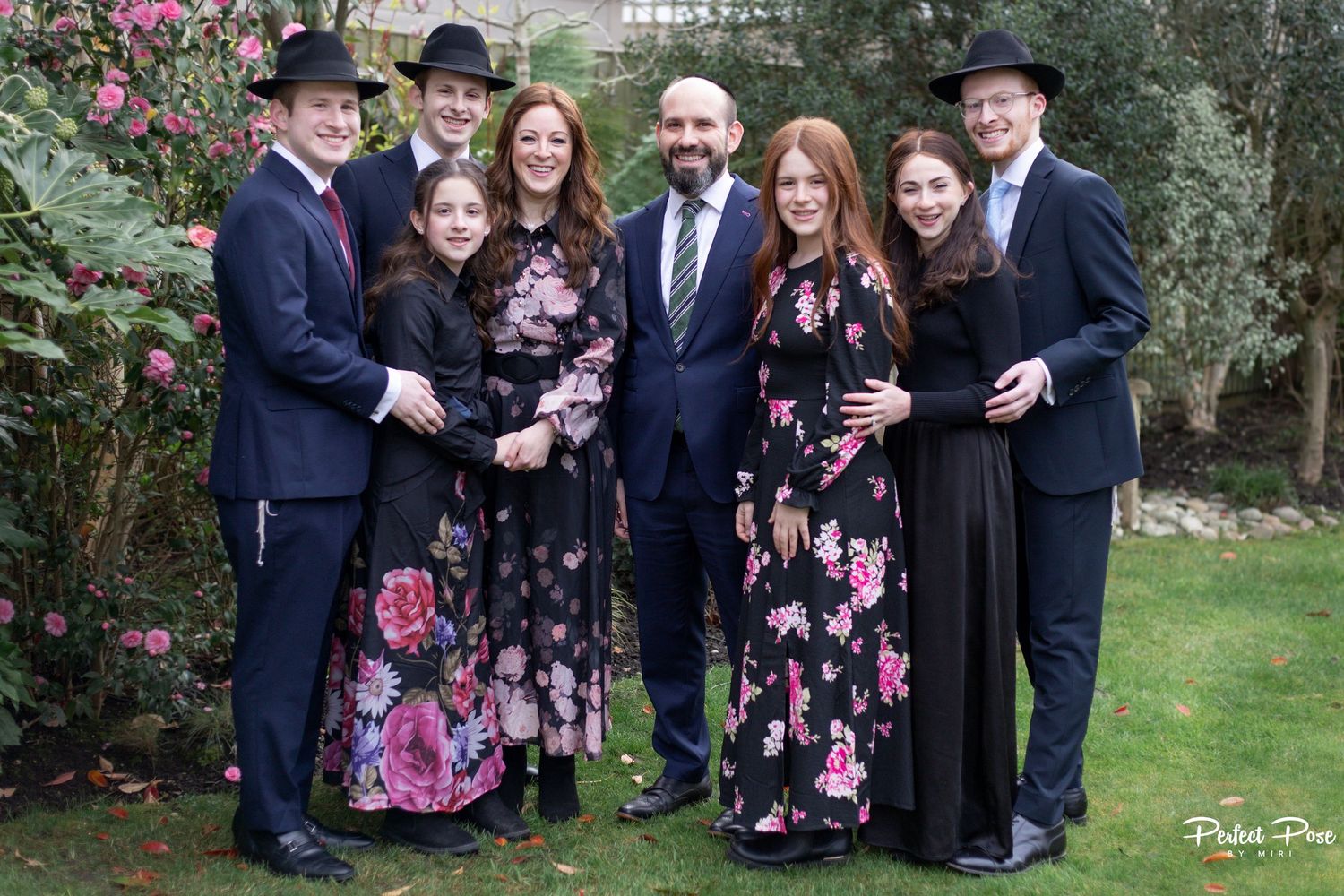 A joyful family group of eight posing outdoors in formal attire, with floral dresses and black suits, in a lush garden.