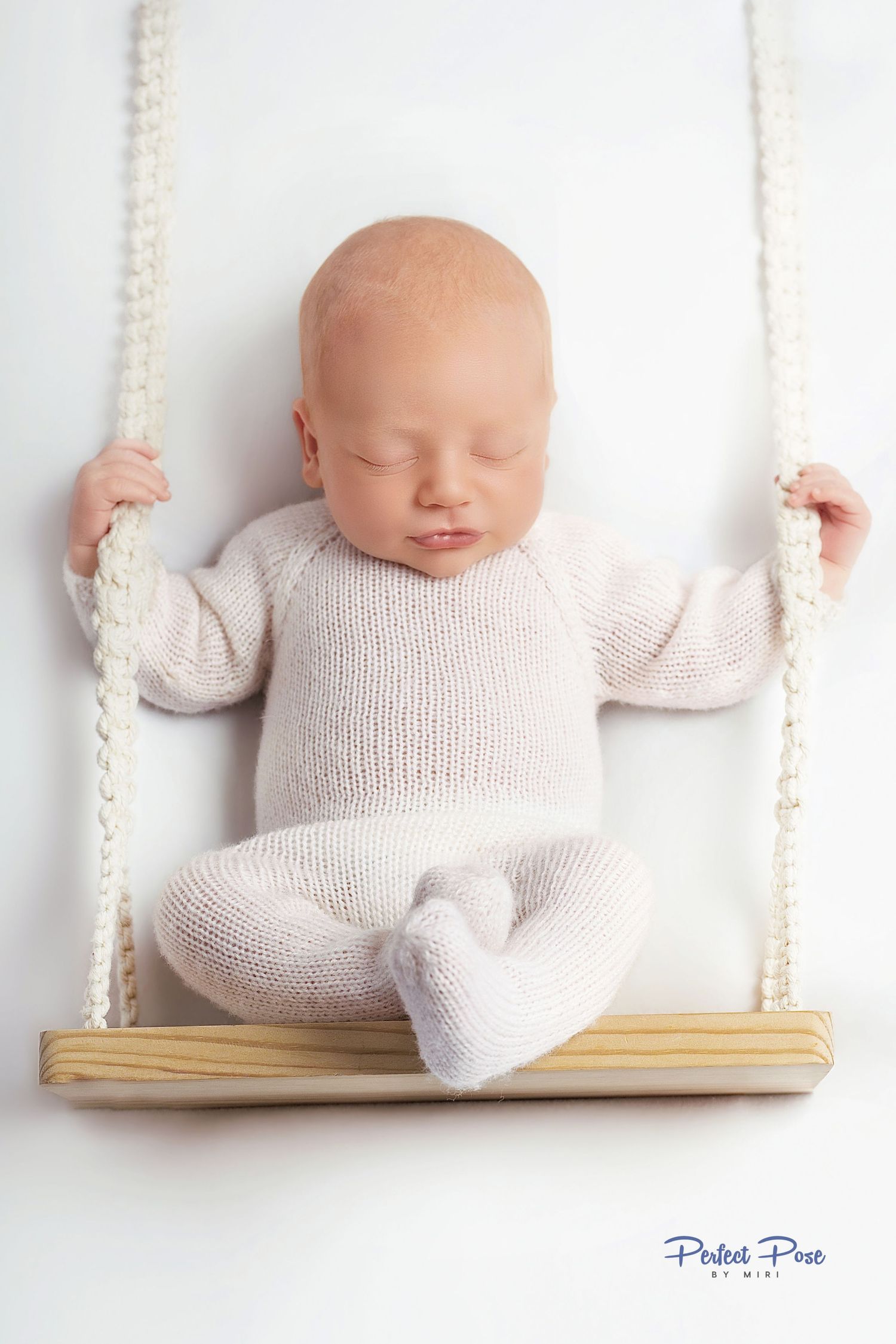 Sleeping newborn baby in cream knit onesie posed on a wooden swing for a professional newborn photography session.