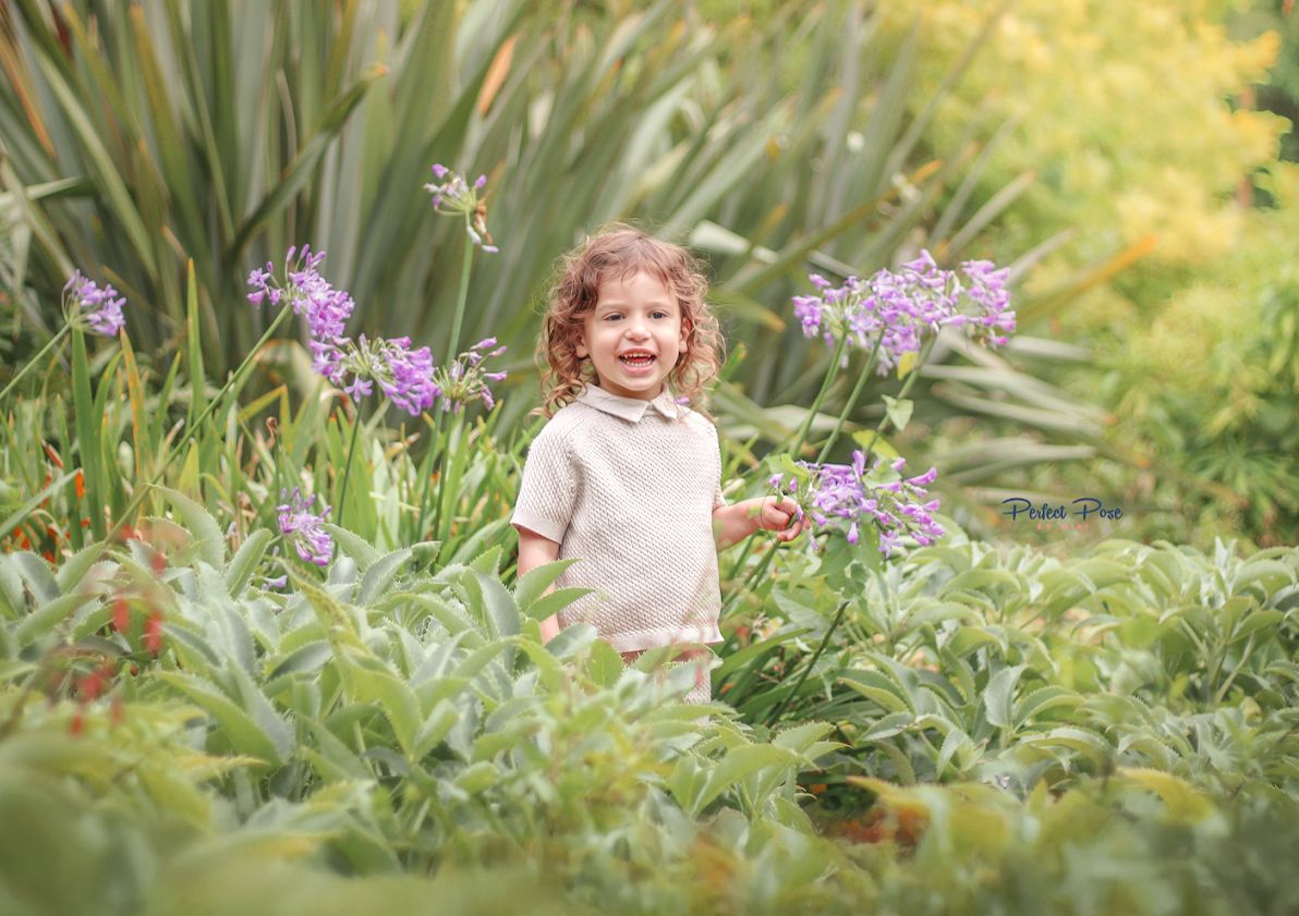 A joyful toddler with curly hair laughs among purple agapanthus flowers in a lush green garden.