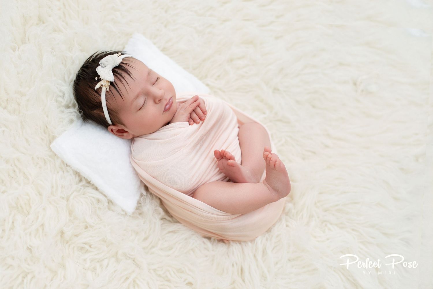 Sleeping newborn baby wrapped in pink swaddle on white fluffy blanket with tiny white bow headband.