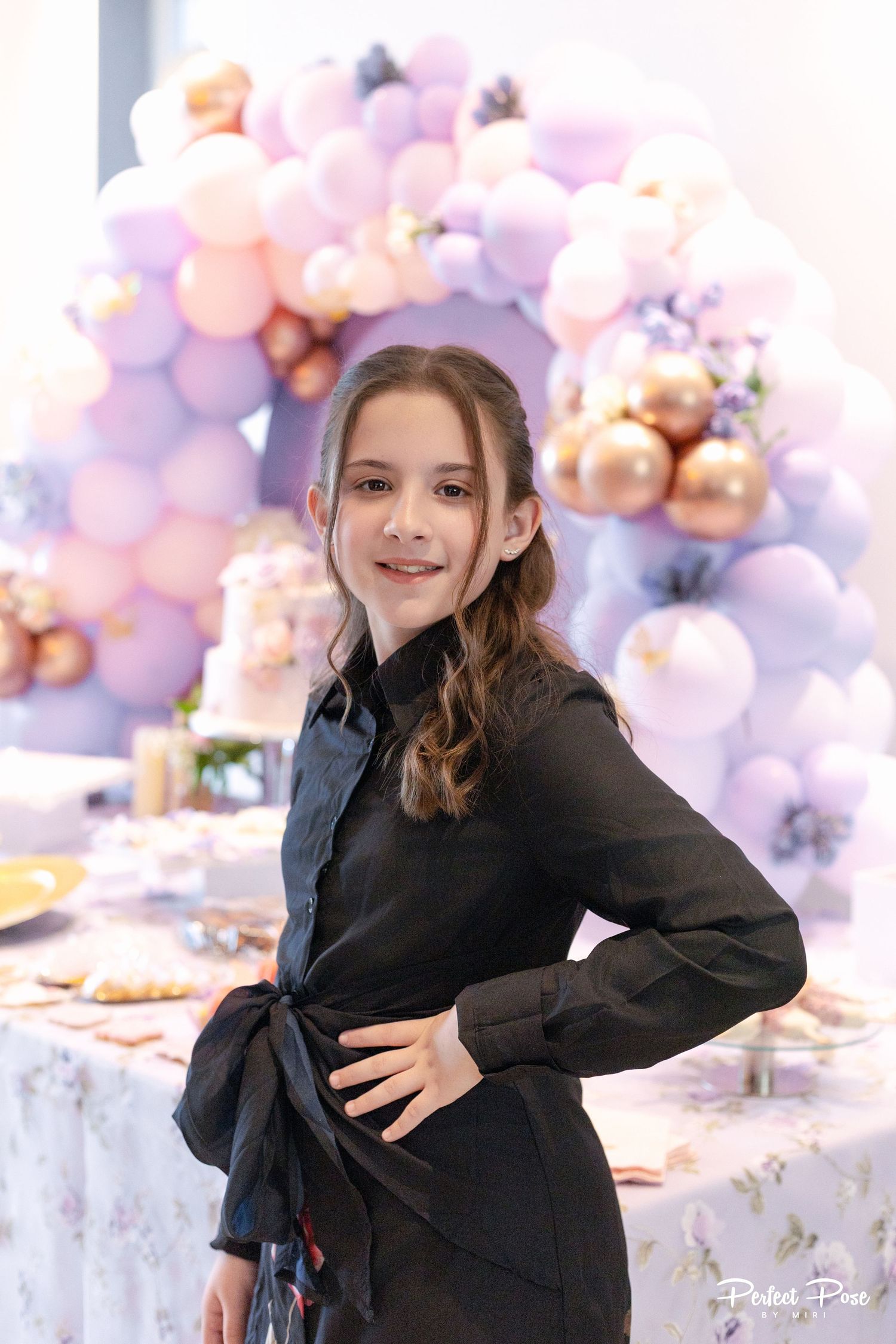 A young girl in a black outfit poses confidently at a birthday party with a pastel balloon arch backdrop.