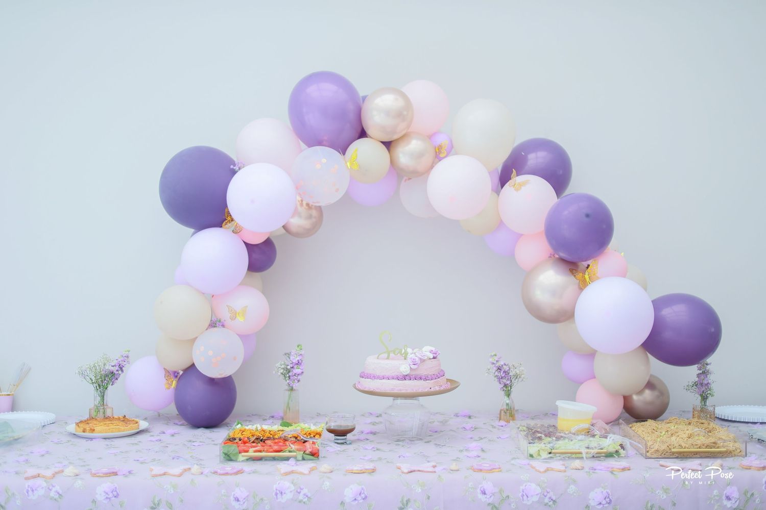 Elegant party dessert table with purple, pink, and gold balloon arch over colorful treats and cake.