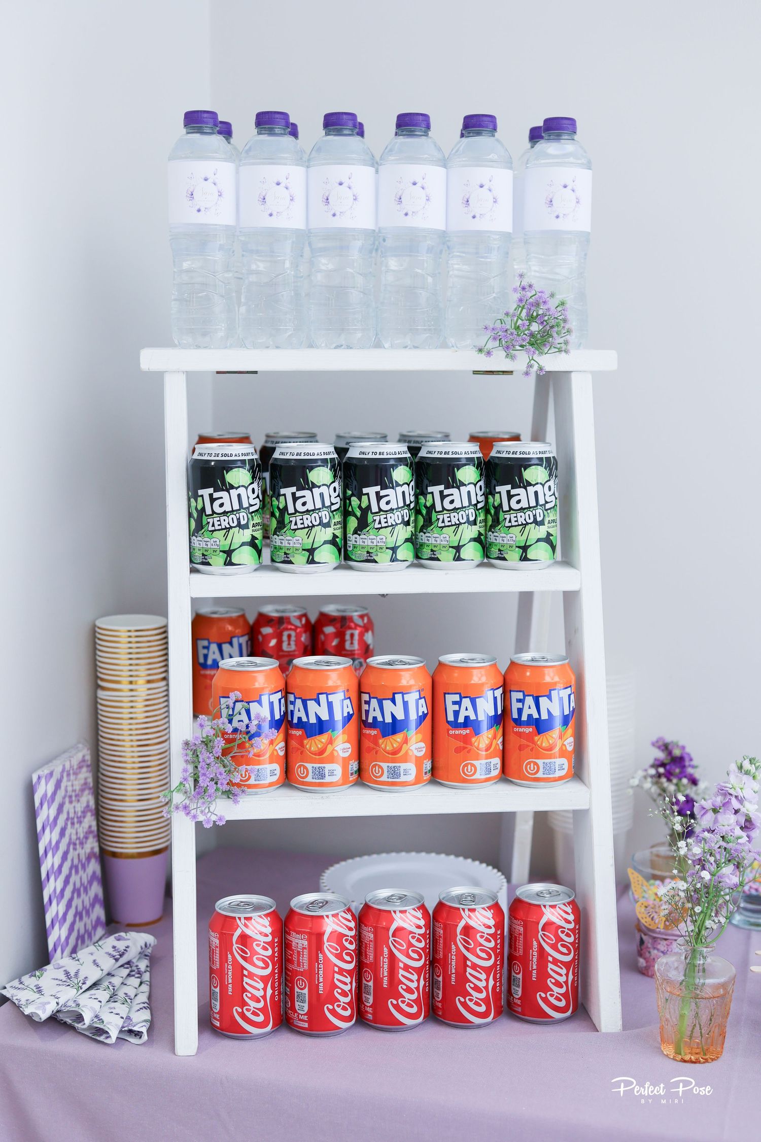 White ladder shelf displaying Coca-Cola, Fanta, Tang cans and water bottles at a party beverage station.
