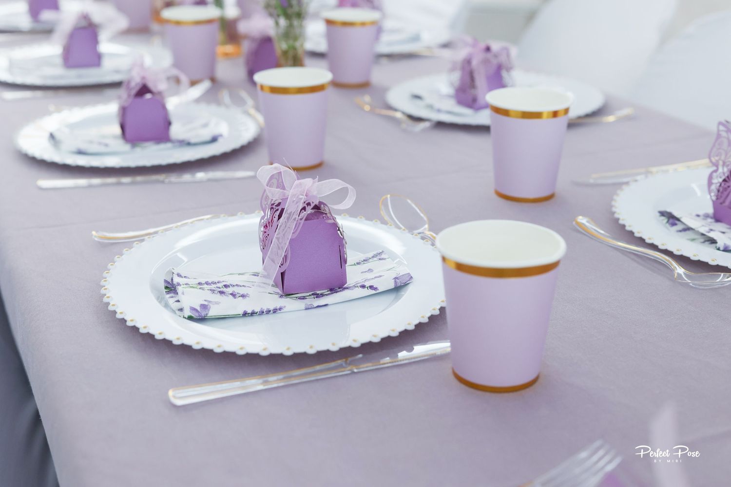 Elegant lavender table setting with purple party favors, gold-rimmed cups, and white scalloped plates for a special occasion.