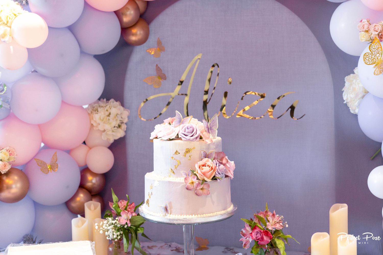 Elegant birthday cake with floral decor and gold name topper surrounded by pink and purple balloon garland.
