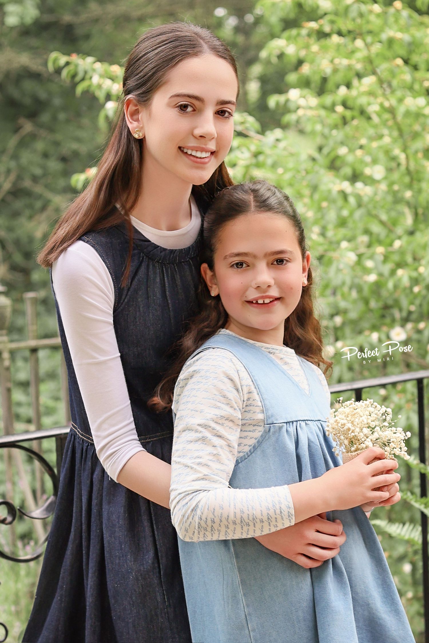 Two girls in denim dresses pose outdoors, the older hugging the younger who holds white wildflowers, with green trees behind.