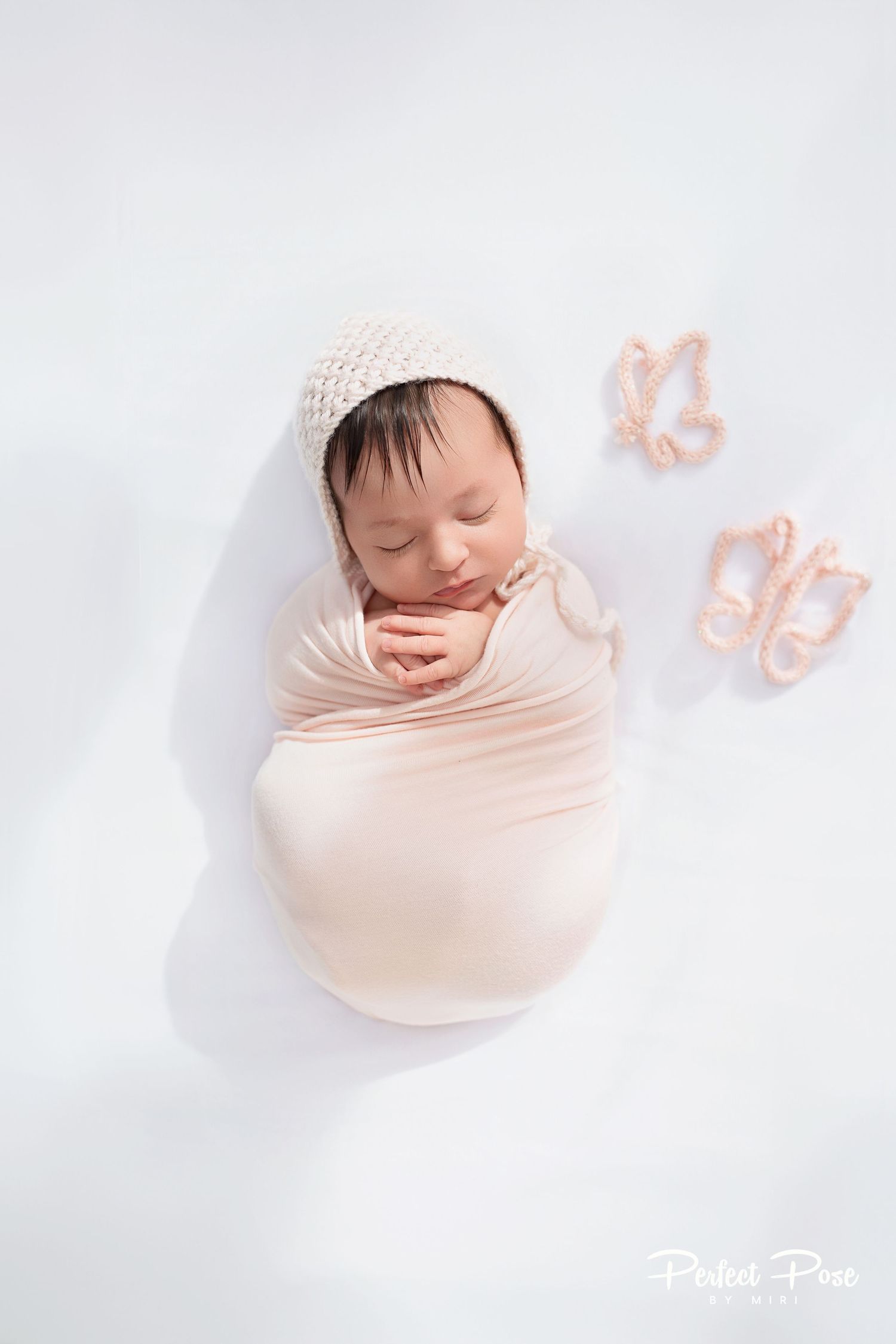 Newborn baby swaddled in pink wrap with knit bonnet, sleeping peacefully beside tiny pink butterfly decorations on white background.