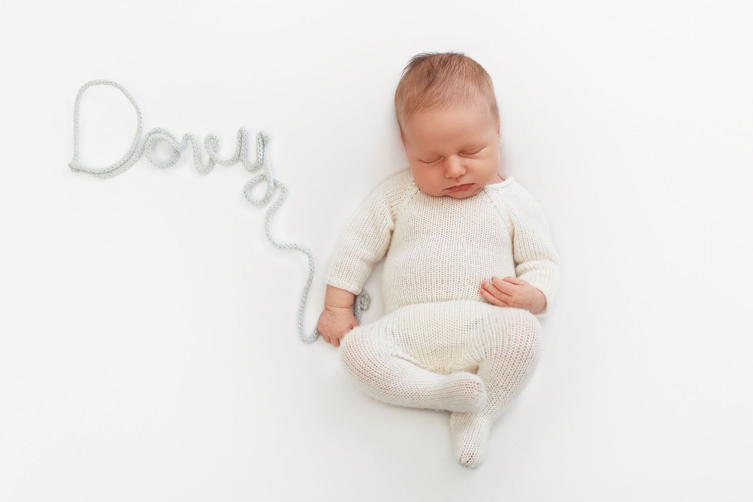 Newborn baby in cream knit outfit lying on white backdrop beside wire name art spelling Donny.
