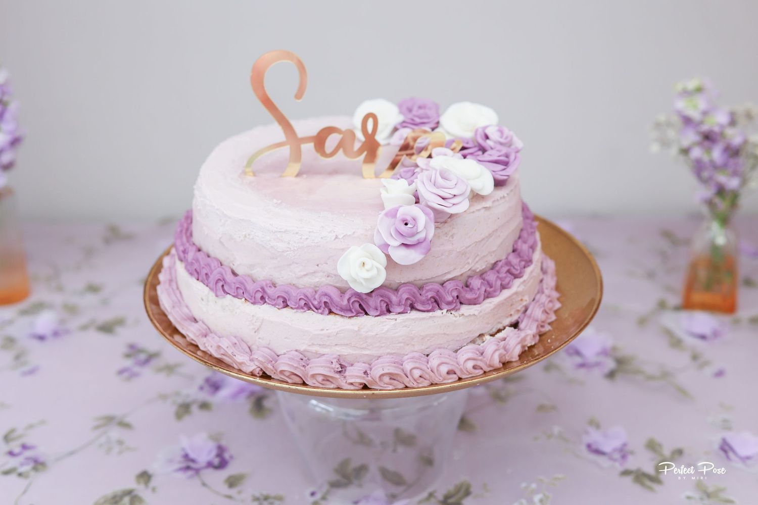 Purple ombre birthday cake with white and lavender roses, gold name topper reading Sara on a floral tablecloth.