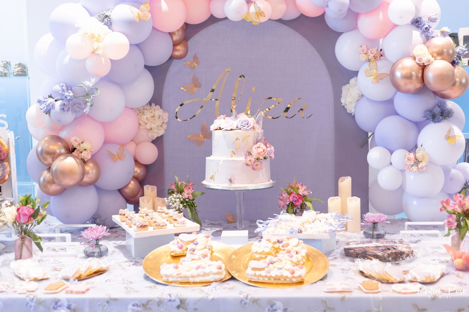 Elegant party dessert table with tiered cake, balloon arch in pink, purple, and gold, and floral decorations.