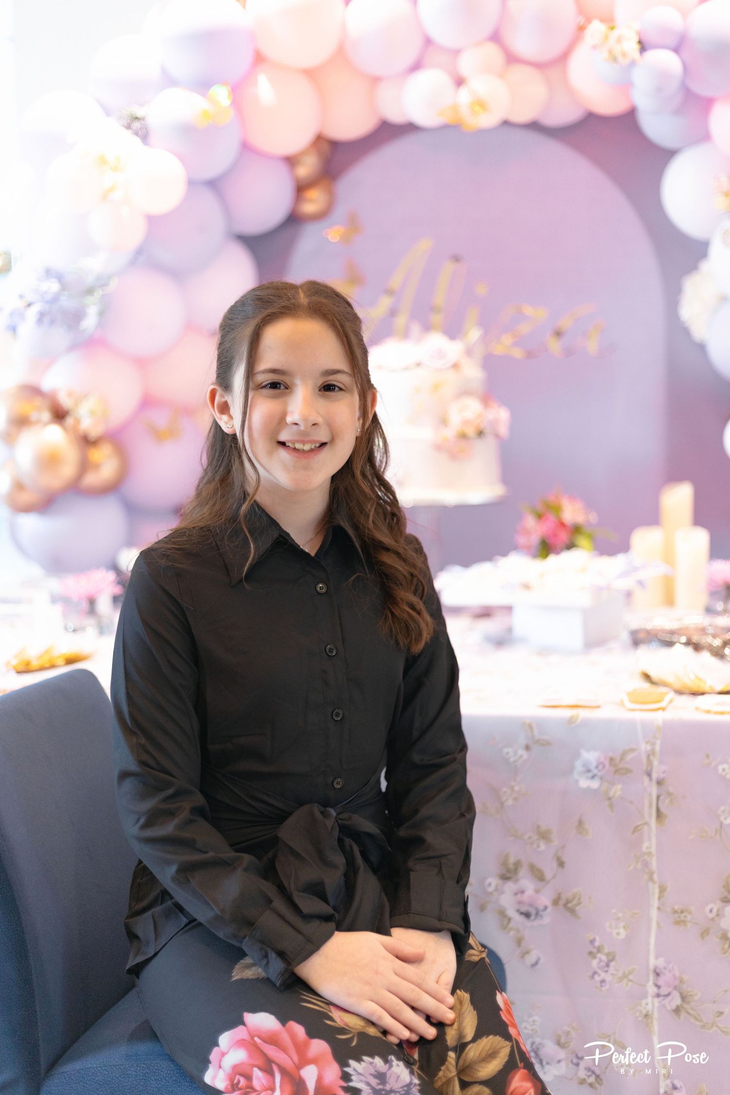 A young girl in a black shirt smiles at a birthday party with pink and purple balloon arch and cake table.