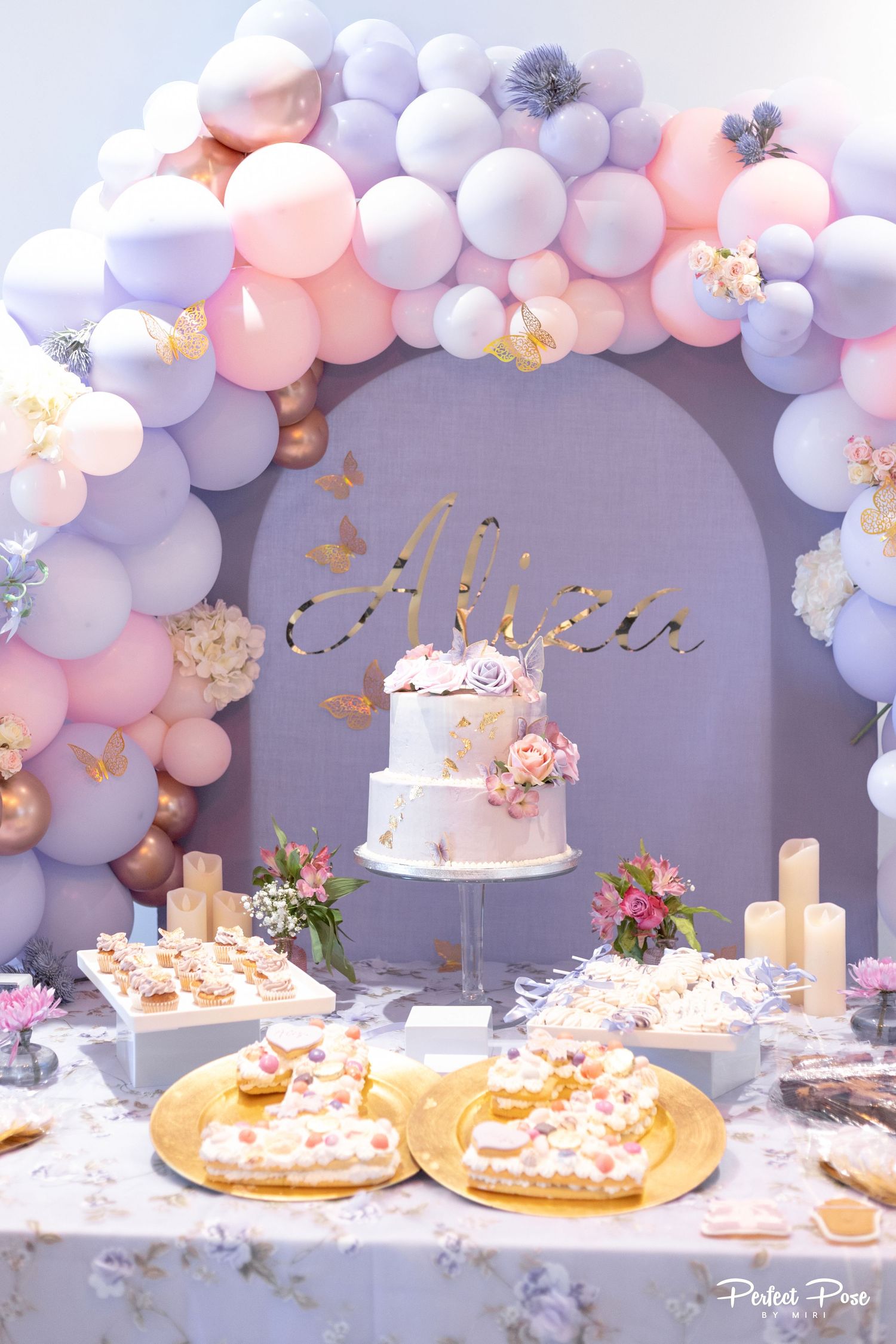 Elegant birthday dessert table with pastel balloon arch, tiered floral cake, and sweet treats in pink, lavender, and gold.