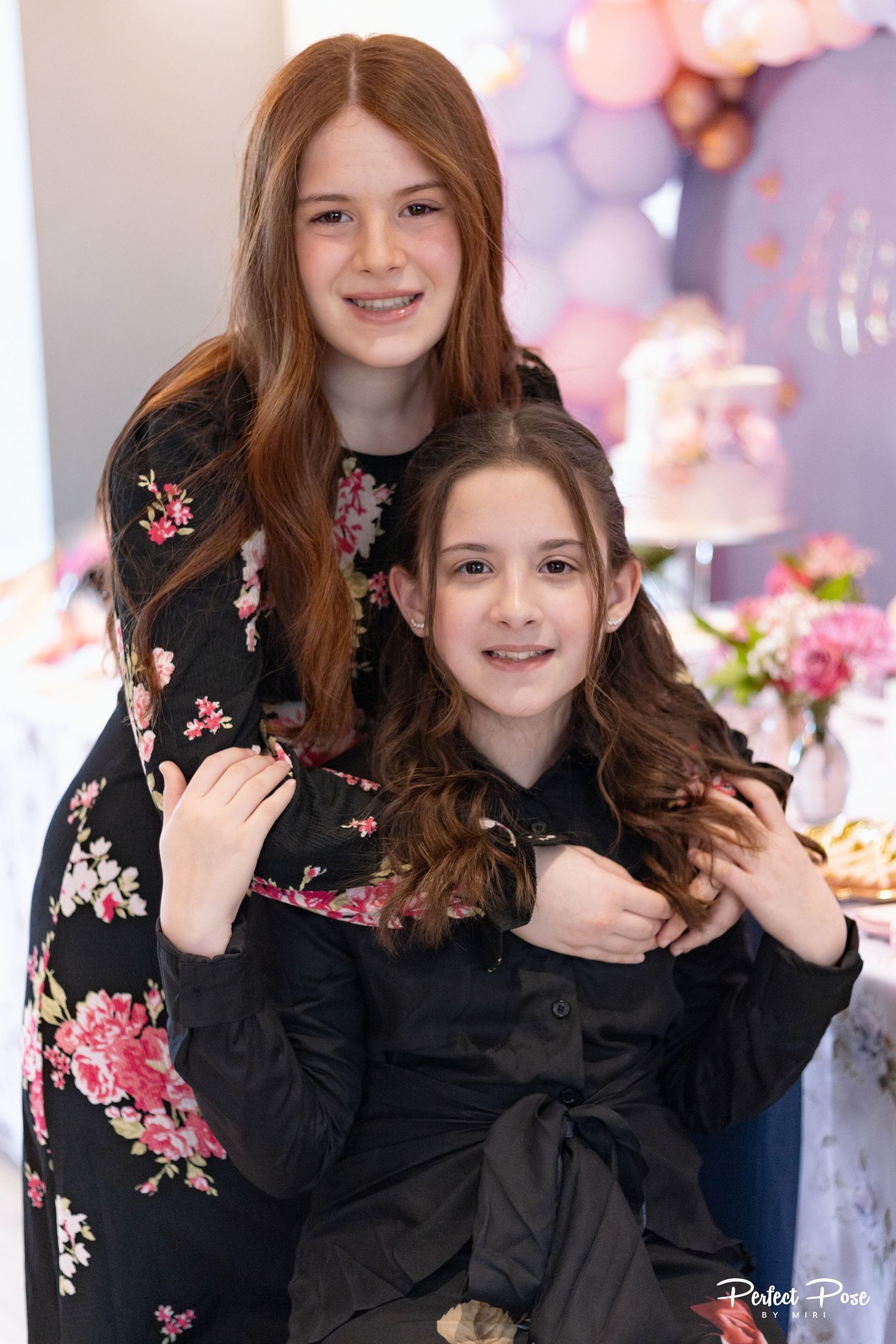 Two young girls in black dresses pose together at a pink and purple party celebration.